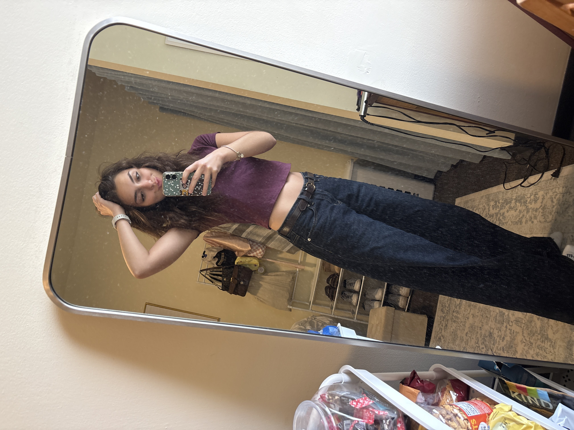 A young woman with curly brown hair is taking a mirror selfie. She is wearing a short-sleeved purple top and dark blue jeans with a brown belt. She holds her phone in her right hand, her left hand resting on her head. The mirror reflects a room interior: shelves with shoes and bags, a rack with snacks, a rug, and a window with blinds. A power strip and connected wires are visible on the wall behind the blinds. The image is taken indoors.