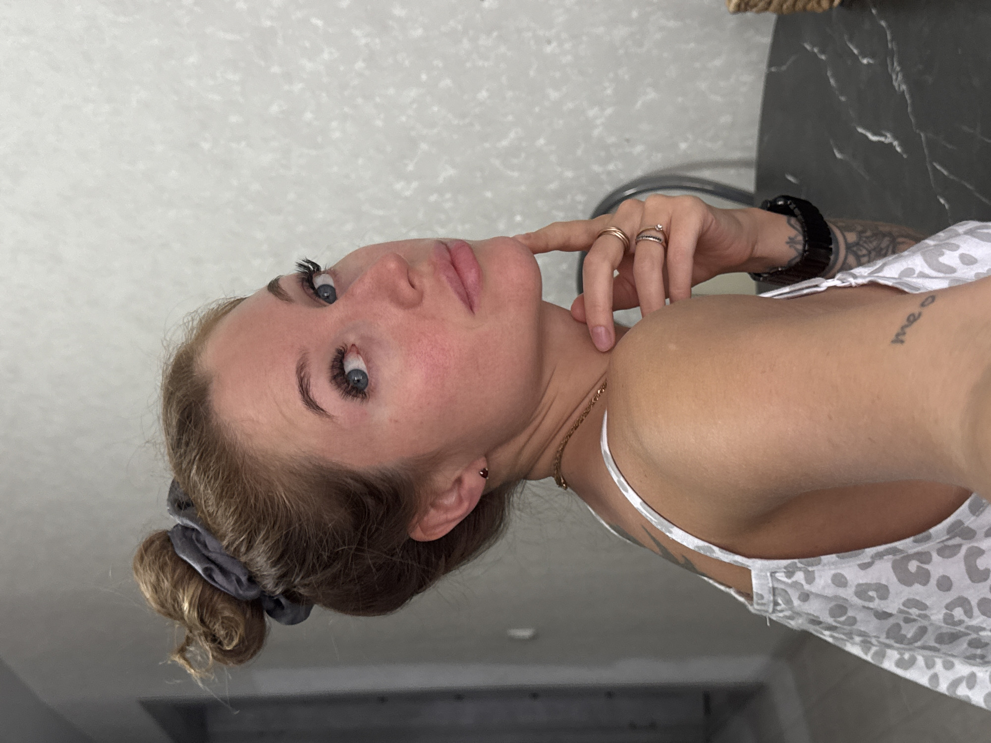 A woman with fair skin and blue eyes is positioned in the center of the image. She wears a white tank top patterned with grey leopard spots. Her light brown hair is pulled back into a low bun secured with a grey scrunchie. She is wearing multiple rings on her left hand and a black bracelet on her left wrist. Tattoos are visible on her left shoulder and inner arm. Her left hand is raised to her face with her fingertips lightly touching her cheek. She has a gold chain necklace. The background consists of a white ceiling and a black marble-like surface. Location: Chaikovsky, Russia is not discernible from this image.