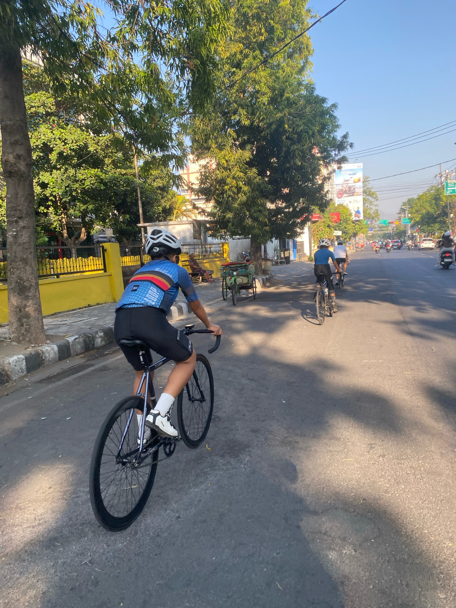 The image depicts a group of cyclists riding on a road in the city of Kediri, Indonesia, during the day. The scene is characterized by a clear sky and bright sunlight casting shadows on the asphalt. In the foreground, a cyclist wearing a blue and red cycling jersey, black shorts, and a white helmet is in motion, with their bicycle's wheels prominently featured. Further down the road, other cyclists are visible, as well as motorbikes and cars, indicating moderate traffic. The surrounding environment includes trees, buildings, and a yellow fence, typical of an urban setting. A traditional two-wheeled cart is parked on the side of the road. The overall atmosphere suggests an outdoor recreational activity taking place within the city.