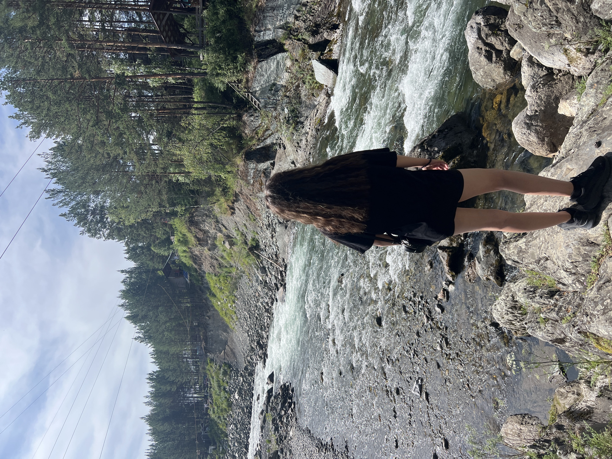 A person with long, dark, wavy hair, facing away from the camera, stands on a rocky bank overlooking a fast-flowing river. The person is wearing a black t-shirt and black shorts, and black shoes. They have a black bag slung over their shoulder. The river is wide and appears to be in a natural, mountainous setting. The banks of the river are lined with rocks and trees. In the background, a dense forest of pine trees covers the slopes of the hills. There are power lines stretching across the sky in the upper left portion of the image, and some wooden structures visible among the trees on the right bank. The sky is partly cloudy.