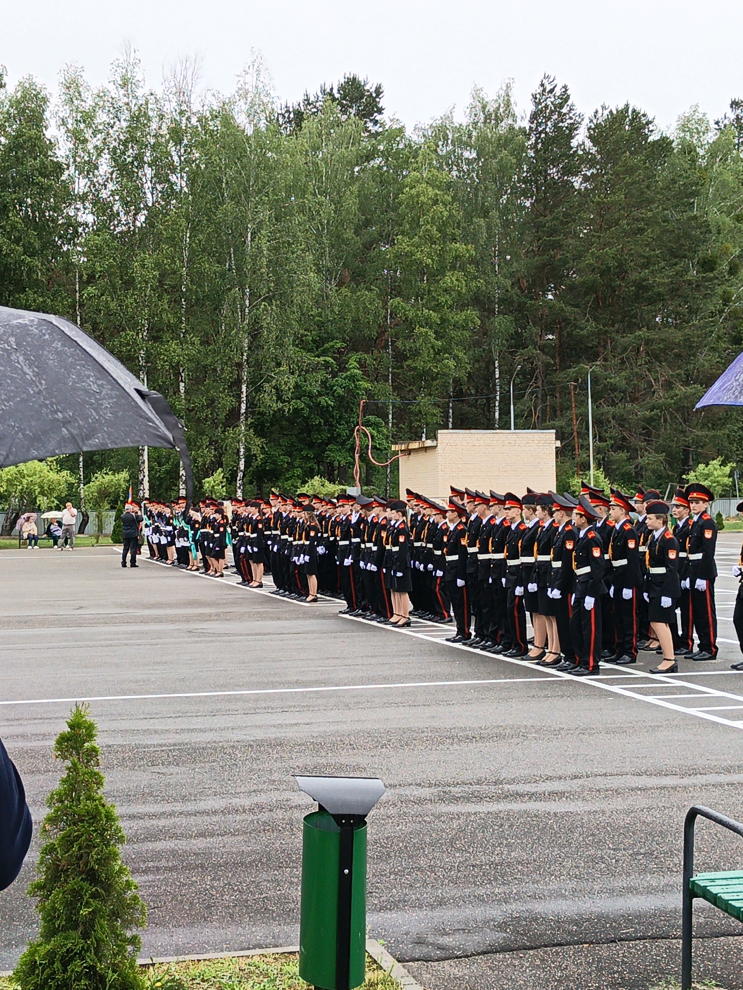 An outdoor scene depicts a uniformed group of individuals, likely students, standing in formation on a paved surface. The individuals are wearing matching black uniforms with red trim and distinctive hats. They are arranged in several rows, facing towards the right side of the frame. In the background, there is a line of trees, suggesting a park or outdoor training area. To the left, a large dark umbrella is partially visible, offering shade. Further left, a few spectators are seated on benches. In the foreground, a small evergreen tree and a green trash receptacle are present. The ground is wet, indicating recent rain or damp conditions. The overall atmosphere appears to be formal, possibly a school event or ceremony. The location is identified as Poreccha, Belarus.