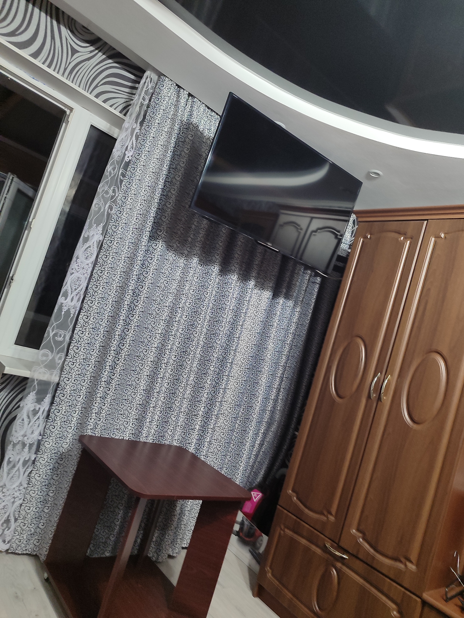 The image depicts the interior of a room, possibly a bedroom or living room, with a distinct architectural style. A large, dark-colored television is mounted on the wall above patterned curtains. To the right, a wooden wardrobe with two doors and ornate detailing stands prominently. In the foreground, a small, dark red wooden table with a curved top is visible. The walls are adorned with a patterned wallpaper, and a white window frame is partially seen on the left. The ceiling features a curved design with a glossy, dark finish in the upper section. The overall impression is that of a furnished living space. The location is identified as Mykolaiv, Ukraine.