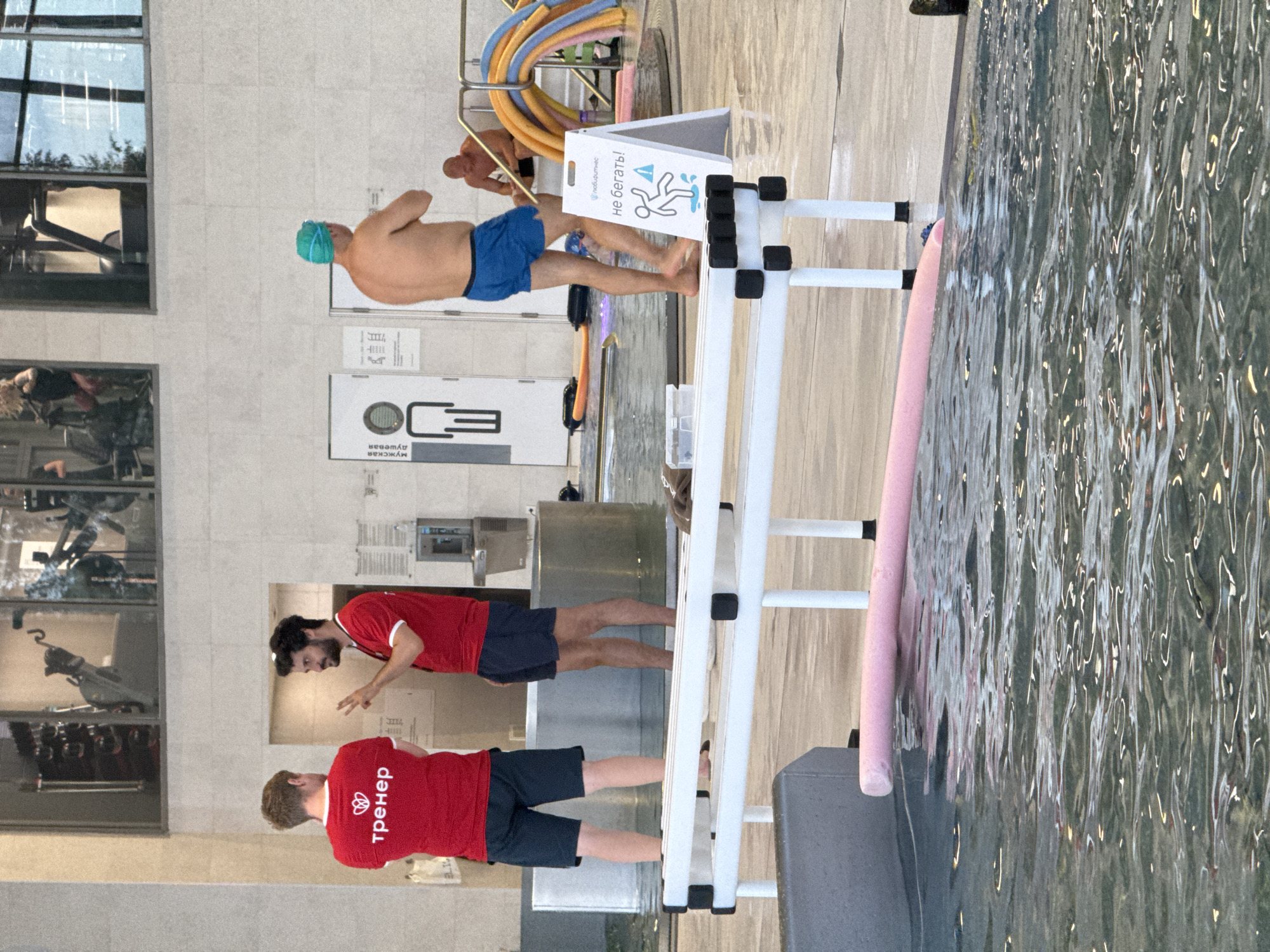 This image depicts an indoor swimming pool facility, likely in Sovkhoza imeni Lenina, Russia. The scene captures several people in and around the pool area.
In the foreground, two individuals wearing red t-shirts with the word "тренер" (trainer) visible on the back of one are interacting. One is gesturing with his hands as if explaining something, while the other is facing away from the camera. Both are wearing dark shorts.
Further back, a man with a blue swimming cap is standing in the shallow end of the pool, wearing blue swim trunks. He appears to be engaged in some activity within the pool, possibly training. Behind him, a sign is propped up near the pool edge with an image of a person slipping and text that reads "не бегать!" (do not run!).
The pool area has tiled walls and a wooden-like floor. A white metal structure with black handgrips and black cubes is visible, likely a pool ladder or a support for equipment. A pink pool noodle rests on the edge of the pool. In the background, through windows, exercise equipment like treadmills can be seen in what appears to be a gym area. The lighting suggests it is daytime, with artificial lights also illuminating the pool area. The overall atmosphere seems to be active, with people engaged in swimming or related activities.