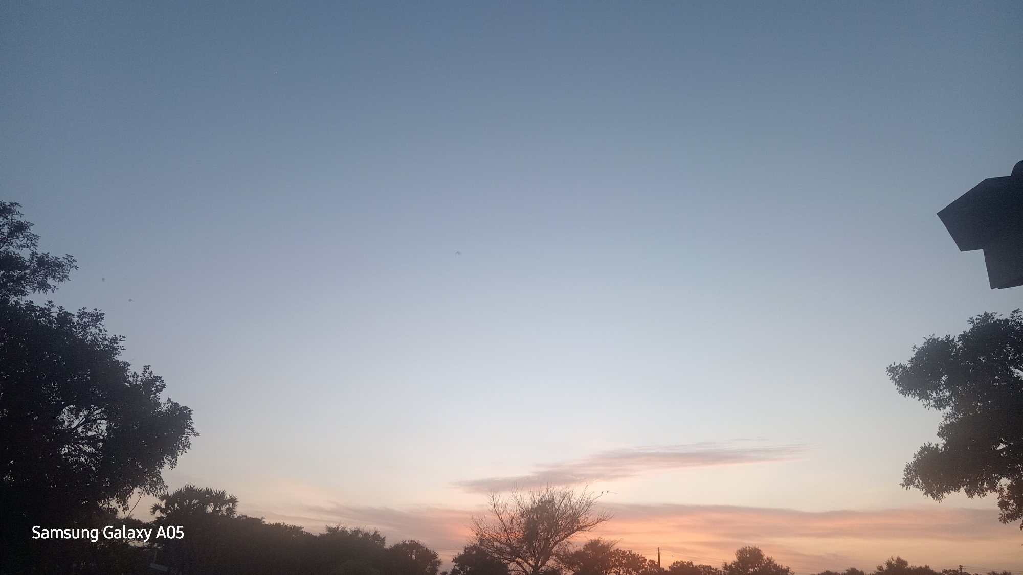 The image depicts a serene sky at dusk, with the horizon illuminated by soft hues of orange and pink. Silhouetted against this twilight sky are the dark shapes of trees and a building. The Samsung Galaxy A05 watermark is visible in the bottom left corner, indicating the device used to capture the photograph. The scene appears to be set in Ribina East, Nigeria, a location implied by the provided context, though the image itself does not contain explicit geographical markers. The overall atmosphere is tranquil and atmospheric, capturing the quiet transition from day to night.