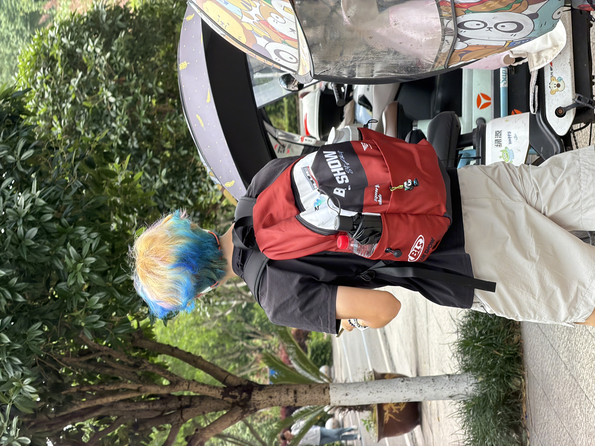 The image depicts a person with dyed blonde and blue hair, wearing a black t-shirt, beige shorts, and a red backpack with the branding "B. SHOW" and "BLS Sport Club." The person is standing next to an electric tricycle or scooter with a sunshade featuring cartoon characters. A water bottle is visible in the backpack. The setting appears to be an outdoor urban environment, with trees and pavement visible. The presence of a plant in a pot with Chinese-style ornamentation along with the style of the scooter suggests the location is likely in the Chongqing High-tech Zone, China. The scooter is parked on what appears to be a paved path.