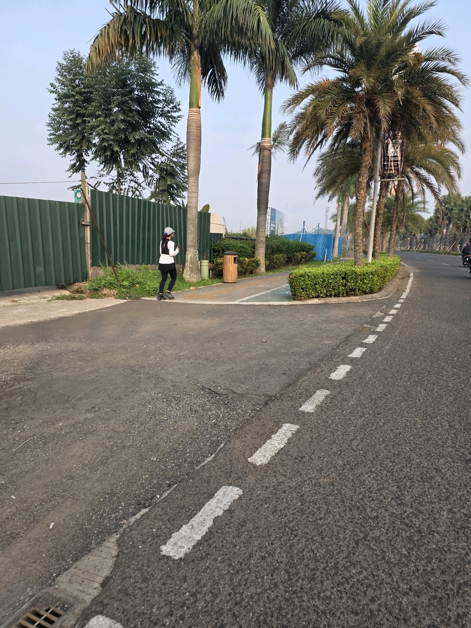 The image shows a street scene in Bojongsoang, Indonesia. A person is jogging on a paved path next to a road lined with palm trees. A green corrugated fence runs along the left side of the path, behind which are some trees and a building. There is a wooden trash bin on the path. The road has white dashed lines marking lanes. In the background, more palm trees and some construction can be seen. The sky is clear and the lighting suggests it is daytime.