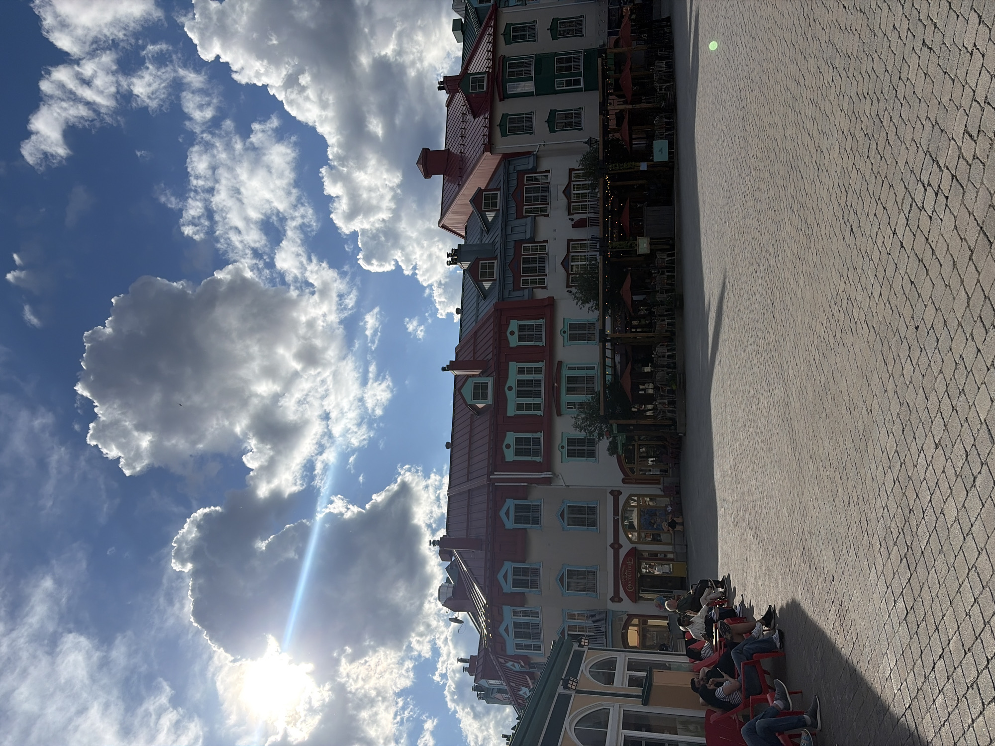 The image depicts a sunny day in the pedestrian village of Mont-Tremblant, Canada. In the foreground, a paved square with interlocking bricks is bathed in sunlight, casting long shadows. A group of people are seated on red chairs, facing the buildings. The buildings, characteristic of the village, feature colorful facades with red roofs, dormer windows, and balconies. Some of the buildings house shops and restaurants with outdoor seating areas shaded by umbrellas. In the background, a bright blue sky is dotted with fluffy white and grey clouds, with the sun shining through, creating a lens flare effect. The overall scene conveys a relaxed, summer atmosphere in a vibrant tourist destination.