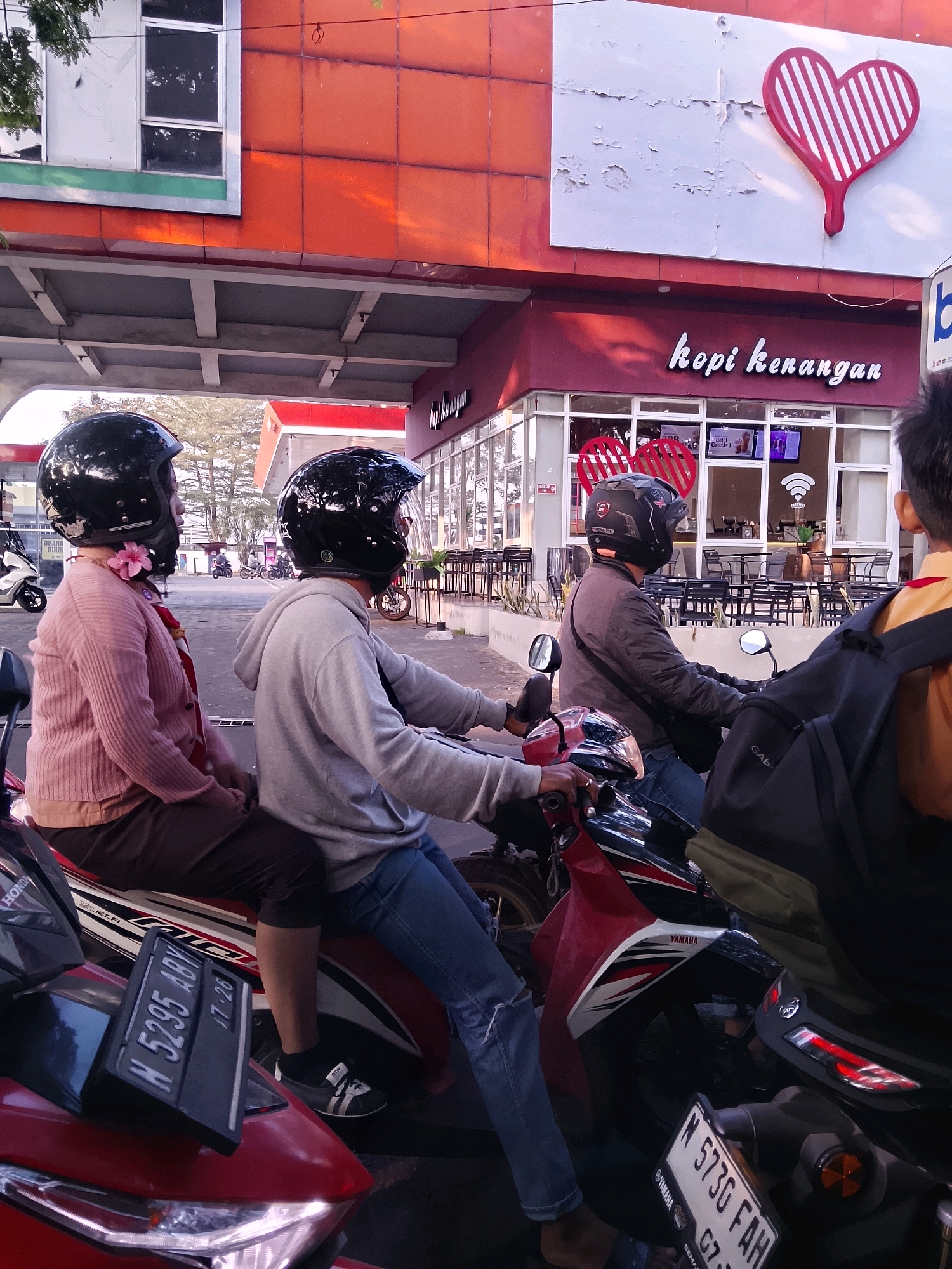This image captures a street scene in Malang, Indonesia, likely outside a "Kopi Kenangan" coffee shop, as indicated by the prominent signage and logo. The foreground features several motorcycles, with two in the center being the primary subjects. On the left motorcycle, a person wearing a pink knitted sweater and a pink flower accessory is seated as a passenger. Next to them, a person in a grey hoodie and blue jeans is driving. Both are wearing black motorcycle helmets. The motorcycle they are on is red and white with visible "JET-FI" and "YAMAHA" branding. A license plate displaying "N 5295 ABY 17-26" is attached to the front of this motorcycle.
In the background, the facade of the Kopi Kenangan shop is visible. It has a modern design with orange and red panels on the upper level and large windows on the ground floor, revealing tables and chairs inside. A large, stylized red heart logo is mounted on the upper facade. The street appears to be moderately busy, with other motorcycles and people visible in the distance, suggesting a typical urban environment. The lighting suggests it is daytime, possibly late afternoon, given the warm sunlight casting shadows. Another motorcycle with a license plate "N 5730 FAH" is visible in the right foreground.
