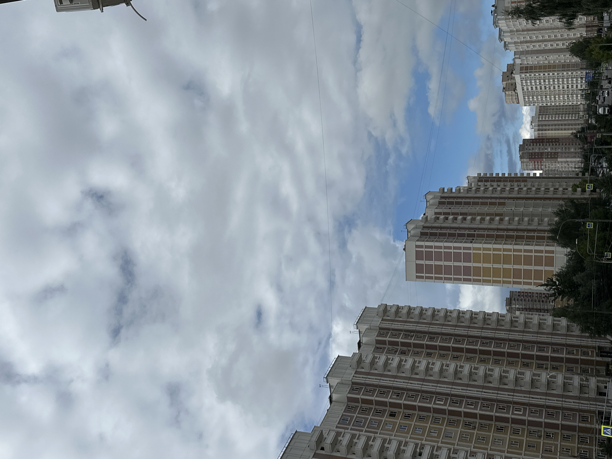 A medium-shot vertical photograph captures a street scene in Podolsk, Russia, under a cloudy sky. The composition is dominated by tall residential buildings that extend upwards, their facades a mix of beige, brown, and white with numerous balconies and windows. The buildings are arranged along a street where traffic is visible, including cars and a truck. Several streetlights and traffic signals are present, along with pedestrian crossings marked with yellow signs. Lush green trees line the street in the foreground and between the buildings, suggesting a residential area. The sky is filled with a mixture of light and dark clouds, with glimpses of blue sky visible in between. There are no people clearly visible in the frame. The overall impression is that of an overcast day in a modern urban environment.