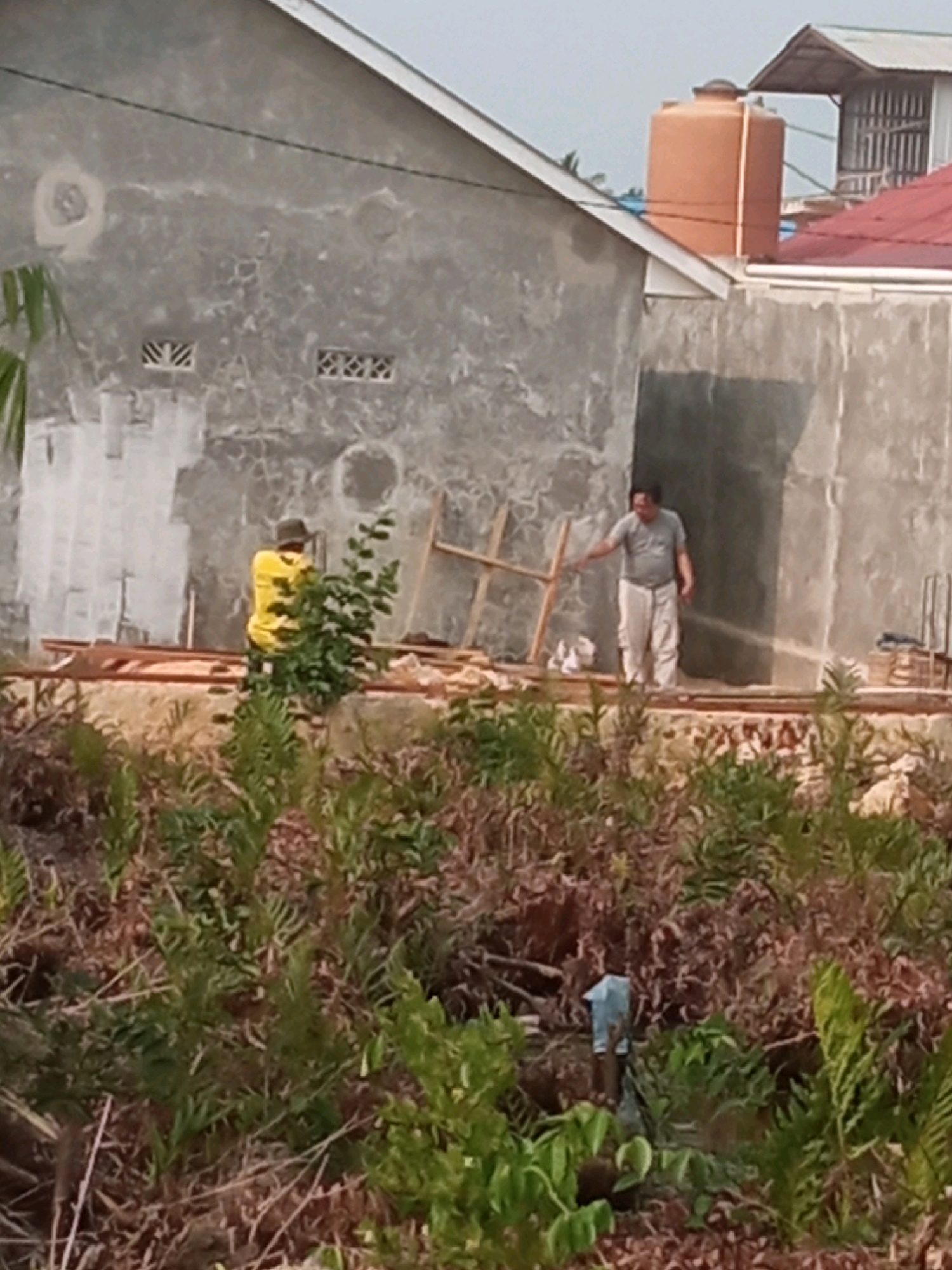 The image depicts an outdoor construction scene in Bontang, Indonesia. In the background, a grey concrete building is under construction, with a large orange water tank visible on its roof. Two men are present on a raised platform in front of the building. One man, wearing a yellow shirt and a hat, stands to the left and appears to be interacting with a small tree. The other man, wearing a grey t-shirt and light-colored pants, is on the right and appears to be spraying water from a hose, possibly to wet the concrete or clear debris. Construction materials, including wooden planks, are visible on the platform. The foreground is filled with dense vegetation and foliage, suggesting a rural or developing area. The sky is overcast.