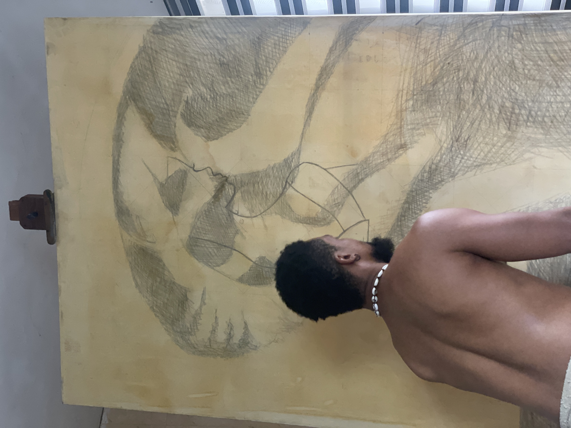 A man, an artist, with dark skin, short dark curly hair, and a beard, is shown shirtless, wearing a white shell necklace, as he intently observes or works on a large, yellowish-brown canvas. The canvas, supported by an easel partially visible on the left, features a partially completed drawing in charcoal or graphite. The artwork is characterized by extensive cross-hatching and bold contour lines, depicting abstract or intertwining shapes. Faint grid lines and the numbers "103" or "101" are subtly visible in the top right quadrant of the canvas. The man's posture conveys deep focus and engagement with his creation. The setting is an indoor space, likely an art studio in Ado, Nigeria, with a striped curtain or blinds visible in the background at the top. The scene captures a moment of intense artistic focus.