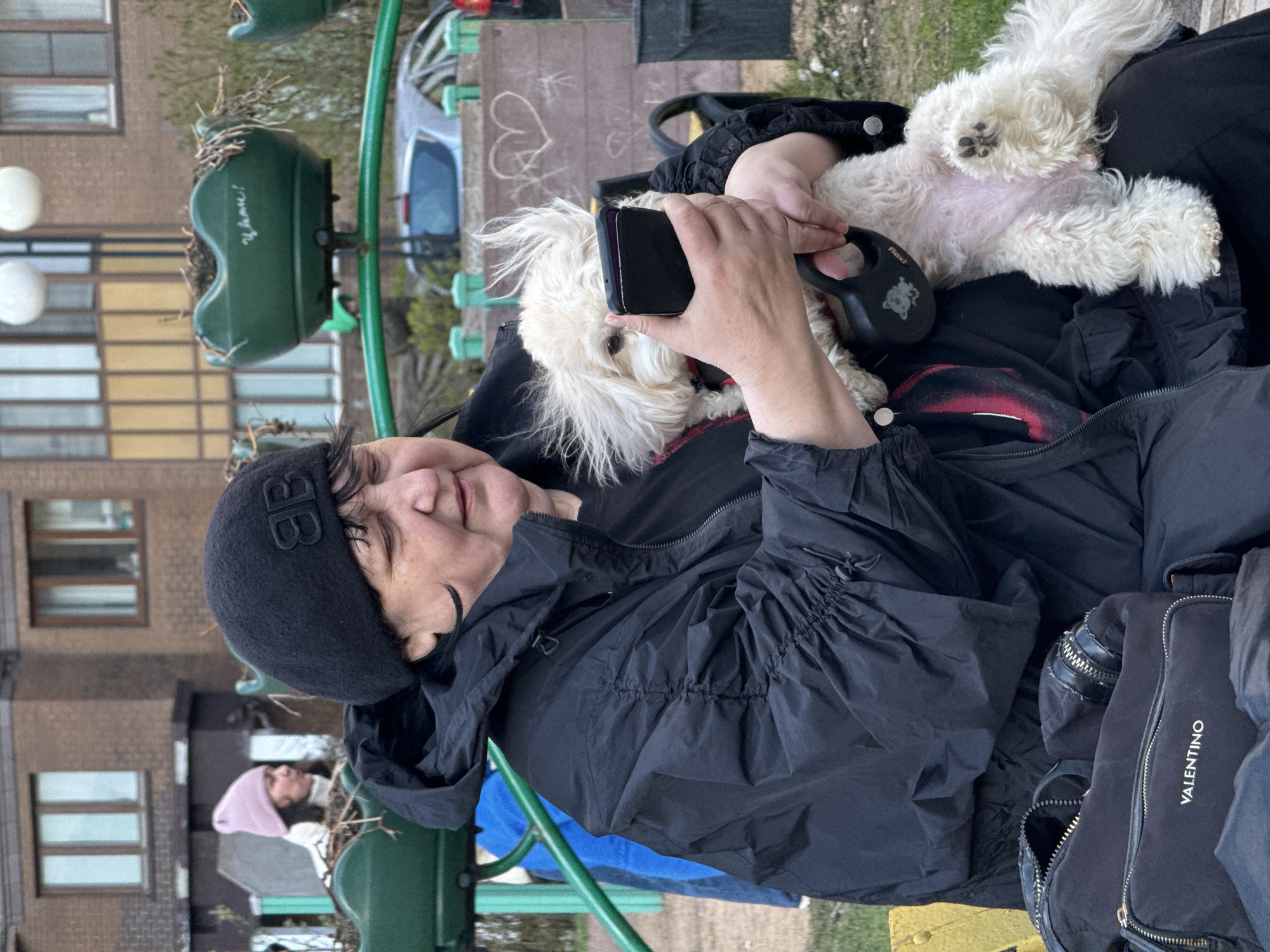 A person is seated outdoors, holding a small, fluffy white dog on their lap in what appears to be an urban or suburban setting, likely in Odintsovo, Russia. The individual, wearing a black beanie with a prominent "BB" logo and a black hooded windbreaker-style jacket, is holding a black smartphone with one hand and a black Flexi brand retractable dog leash with the other.
The small white dog, possibly a Maltese or similar breed, is resting comfortably on its back and side across the person's chest and lap, revealing its light pink belly and a black paw pad. Its fur appears shaggy and dishevelled. The person has a neutral expression, looking slightly upwards and away from the camera.
In the background, residential buildings with multiple windows are visible, along with a partially seen silver car and white round light fixtures. A green metal structure, resembling playground equipment or a bench, extends through the scene. On one of the green elements, the word "Удачи!" (Udači! - Good luck!) is visibly written in white. Another person wearing a pink beanie is partially visible further in the background, near the green structure. A black bag bearing the "VALENTINO" brand name in white text is placed next to the seated person. The outdoor scene suggests overcast or cooler weather, consistent with the subjects' attire.