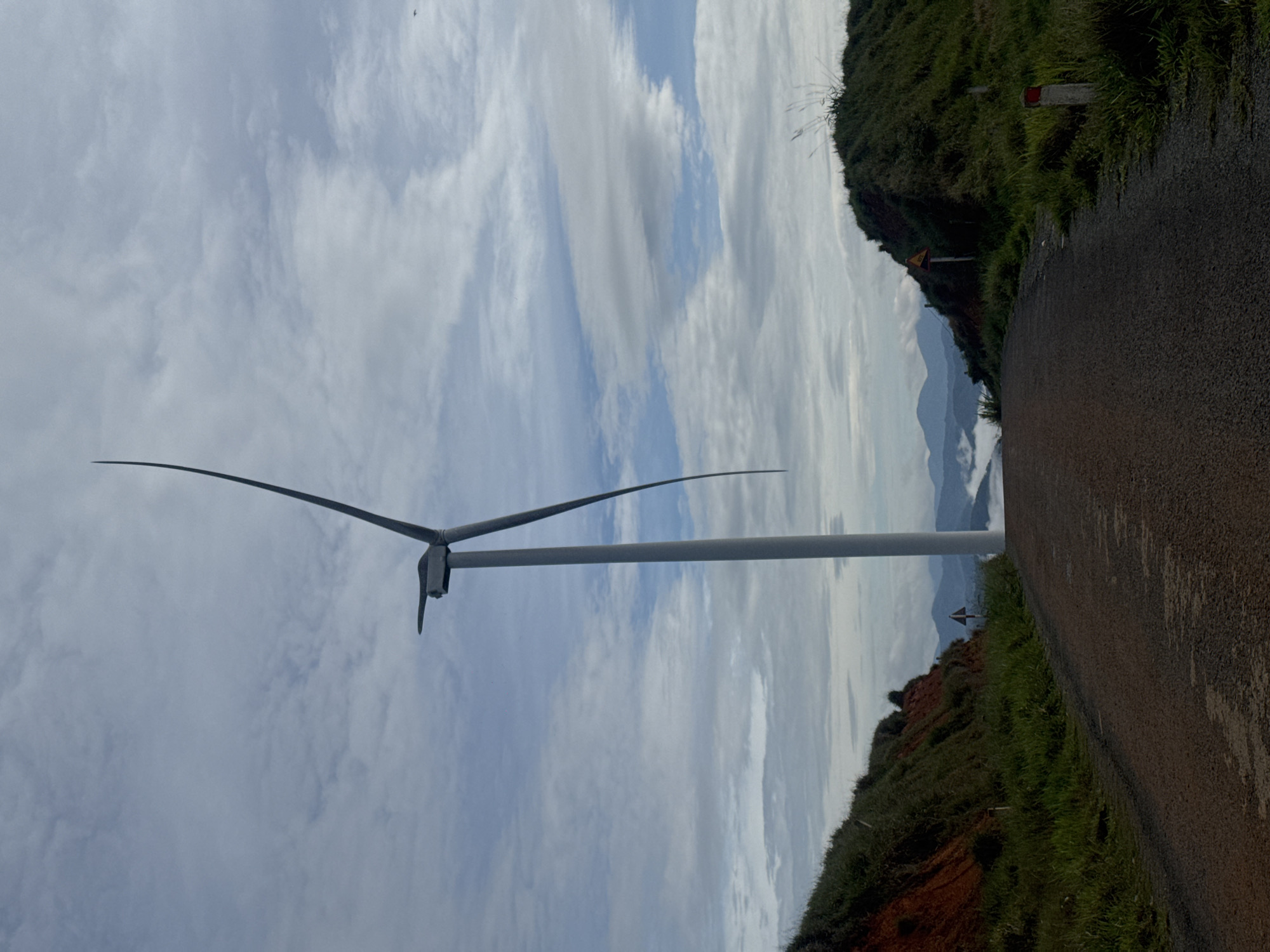 The photograph shows a wind turbine situated near a paved road in Phát Chi, Vietnam. The turbine's blades are prominent against a sky filled with white and gray clouds. The road runs along the edge of a grassy embankment with red soil visible beneath the vegetation. Mountains are visible in the distance, partially obscured by clouds or fog. Traffic signs are present along the roadside.