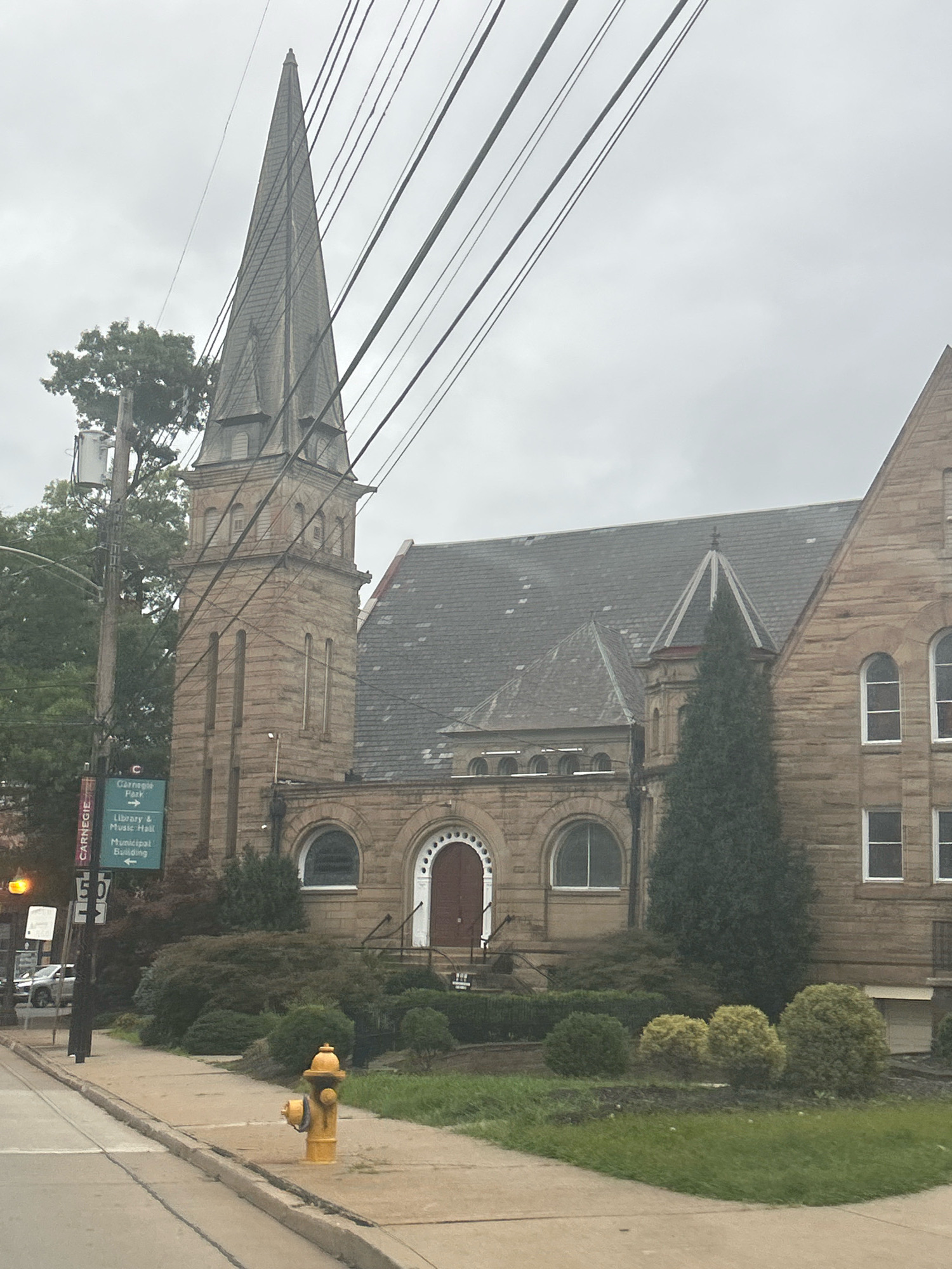 The image shows the Carnegie Library and Music Hall in Carnegie, Pennsylvania, United States of America, on a cloudy day. The prominent feature is the stone church-like building with a tall, pointed steeple and a slate roof. In front of the building, there is a sidewalk and a street with a yellow fire hydrant. A street sign indicates directions to Carnegie Park, Library & Music Hall, and Municipal Building. There are also trees and manicured bushes surrounding the building. Overhead, numerous utility wires crisscross the sky. No people are visible in the image.