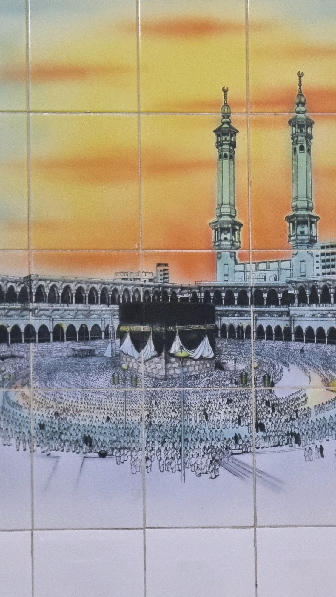 The image is a photograph of a tiled mural depicting the Kaaba in Mecca, Saudi Arabia. The mural shows the Kaaba, a cube-shaped building draped in black cloth, surrounded by a large crowd of people. Two minarets are visible on either side of the complex. The sky in the background is depicted with a gradient of orange and yellow, suggesting a sunset. The mural is installed on a wall, with visible grout lines between the tiles. There is no indication of the location being Puloara, Indonesia, as the scene depicted is an Islamic holy site.