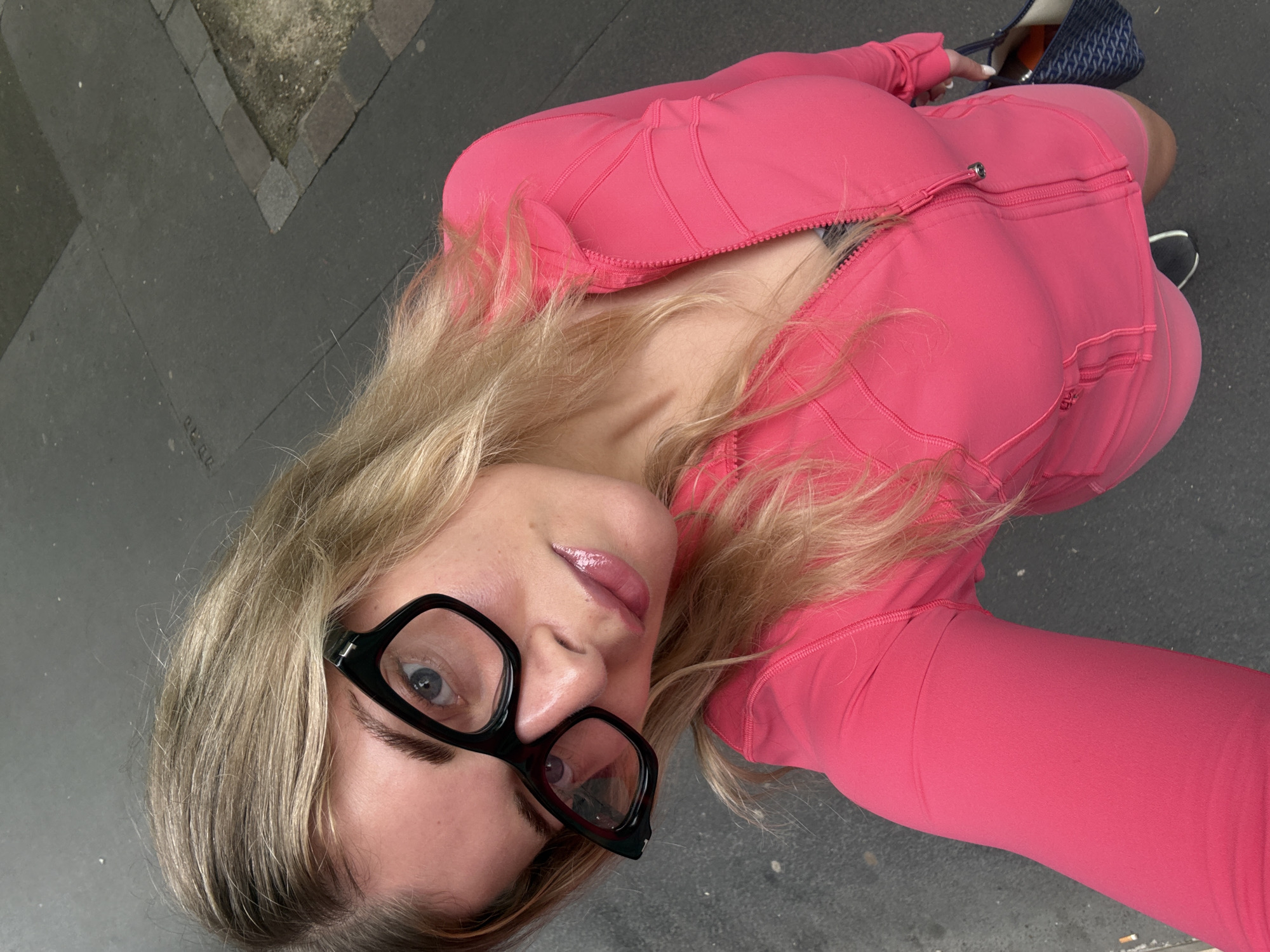 A young woman is seen from a high-angle perspective, likely a selfie, against an urban backdrop. She is wearing large, black-framed glasses and a pink athletic outfit consisting of a zip-up jacket and leggings. Her blond hair is styled loosely, and she has a subtle gloss on her lips. She holds a bag or purse with a patterned fabric. The ground appears to be a concrete surface, suggesting a sidewalk or street. Given the city context and the architecture visible in the background, it is plausible that the photo was taken in Paris, France.