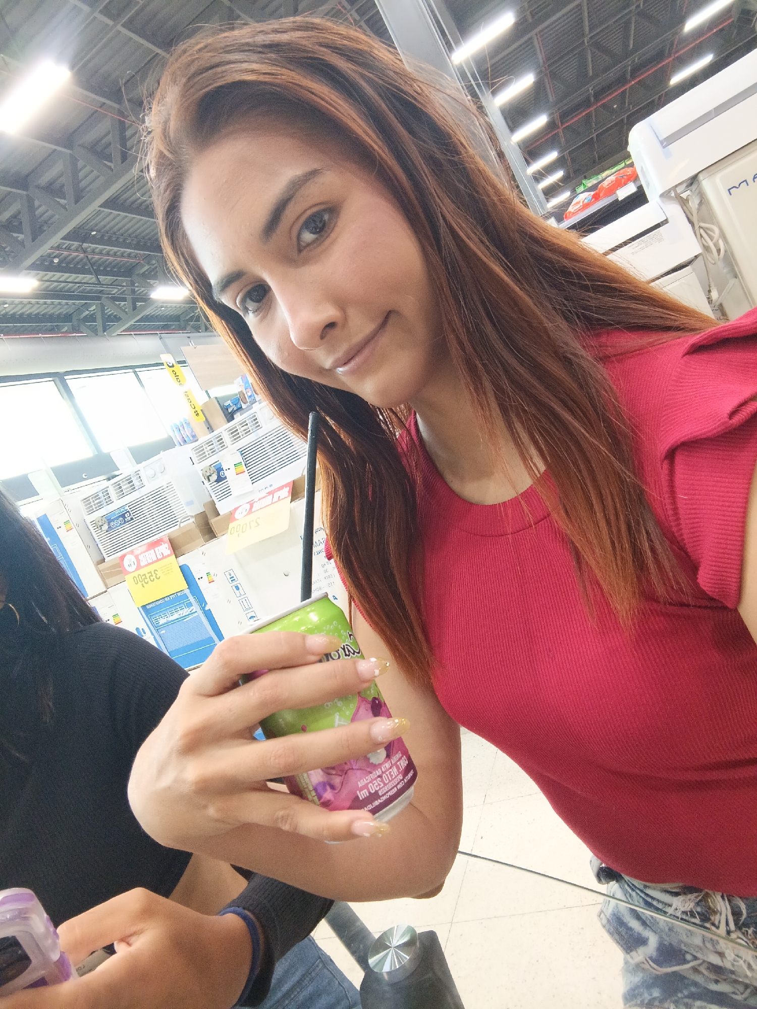 A selfie taken indoors, most likely within a retail environment resembling a department store, given the presence of retail merchandise like air conditioning units in the background. A woman is holding a green can with a straw. The woman has medium-length brown hair and is wearing a red top. Another person is partially visible, with a black shirt and blue jeans.