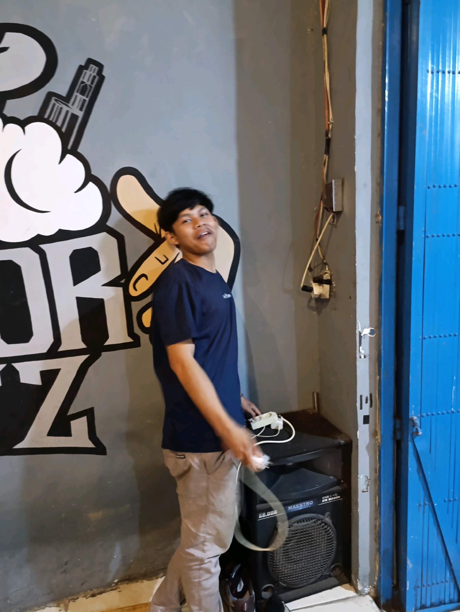 A young man stands in front of a mural in what appears to be an outdoor or semi-outdoor space in Karangsono, Indonesia. The mural depicts stylized clouds and letters, with "POR" and "Z" being partially visible. The man is wearing a navy blue t-shirt and light brown trousers, and he is holding a white power strip with a plug in his right hand, seemingly in the process of plugging it into an electrical outlet mounted on the wall. To his right, a blue metal gate is visible, partially obscuring the background. Below the outlet, a black speaker labeled "MAESTRO GB.000" is present. The overall environment seems to be somewhat industrial or utilitarian, with concrete walls and visible electrical wiring.