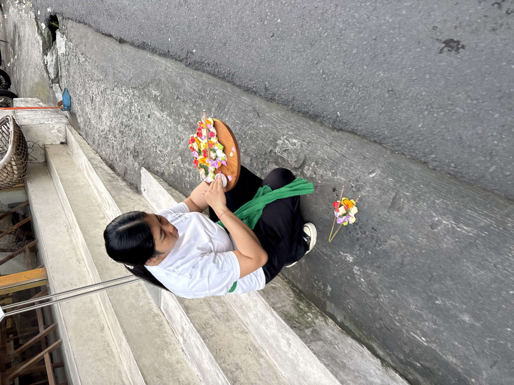 In the city of Ubud District, Indonesia, a young woman with dark hair tied in a ponytail is crouched on the side of a street next to a set of concrete stairs. She is wearing a white t-shirt, black pants, and a green sash tied around her waist. The woman is holding a round wooden tray filled with small, colorful flowers, possibly as an offering. In front of her, on the ground, are two small arrangements of flowers with incense sticks. The street appears to be paved with asphalt, and there is a slight incline. A blue dustpan and a broom are visible near the bottom of the stairs. The scene suggests a cultural practice, possibly related to religious offerings common in Bali.