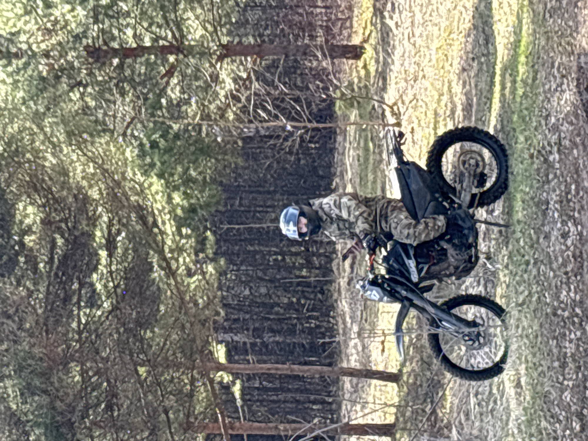 The image shows a person riding a dirt bike through a wooded area. The rider is wearing a helmet and camouflage clothing. The location is not identifiable as Noviy, Russia, based on the visible elements in the image. The rider appears to be navigating a trail or path through the trees. The background suggests a forest or woodland environment with dense vegetation and tree cover.
