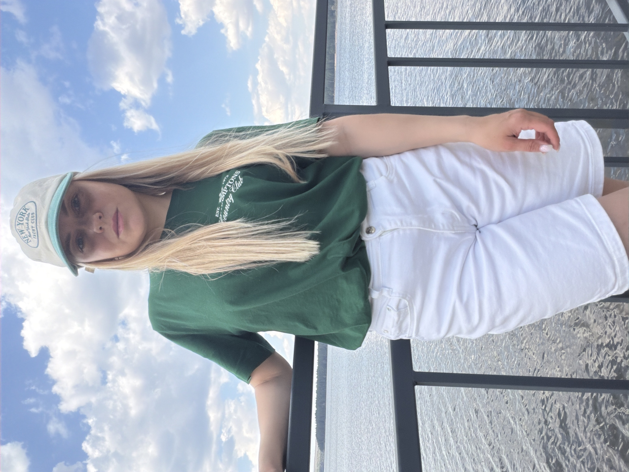 A young woman stands outdoors, leaning against a metal railing. She wears a green t-shirt with a logo and white shorts, and a baseball cap. The background shows a body of water with a cloudy sky above. The location is likely Votkinsk, Russia, given the prompt's context, although specific Votkinsk landmarks are not visible.