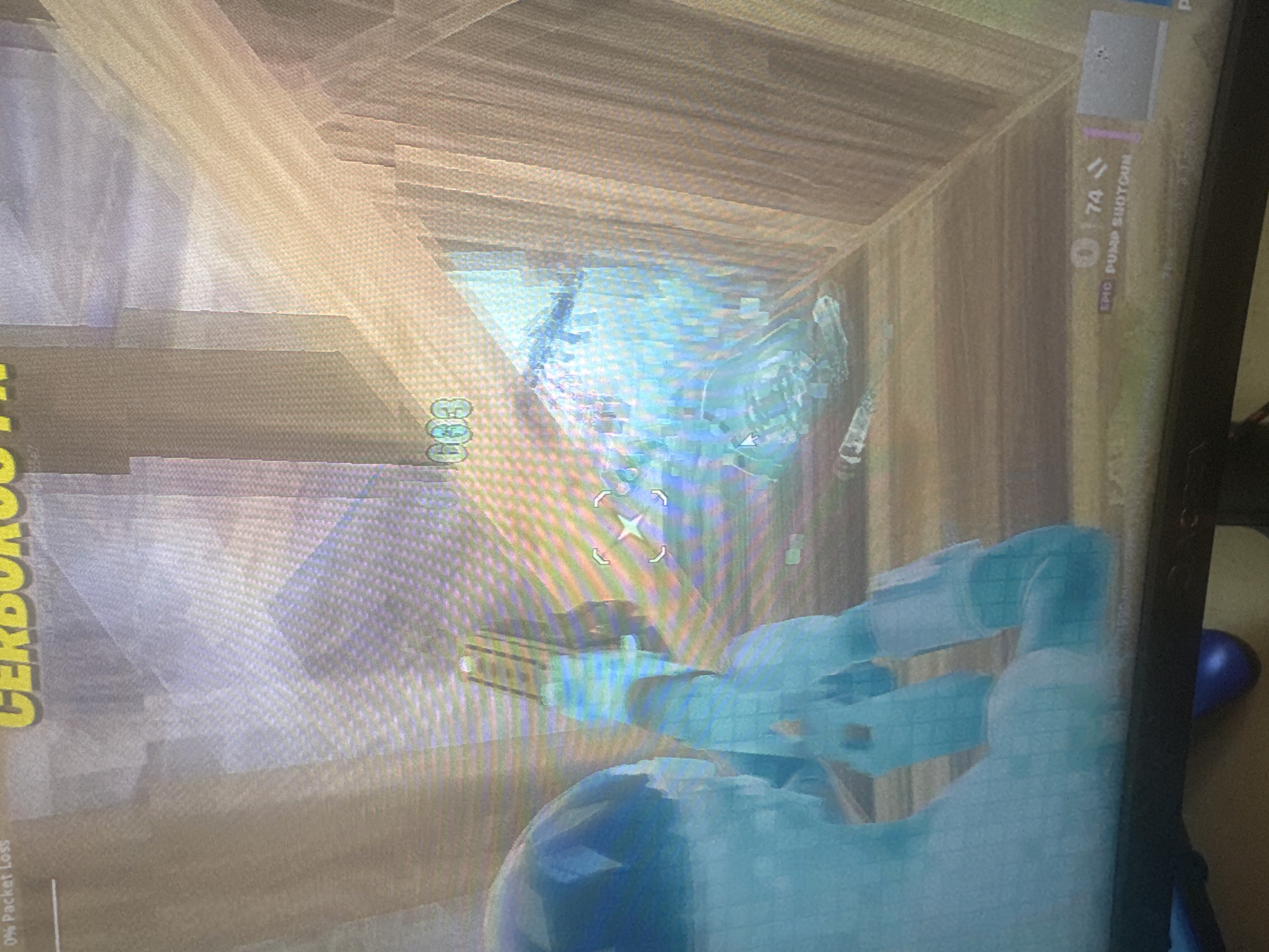 The image shows a first-person perspective of a video game, likely Fortnite, as indicated by the user interface elements. The player character, rendered in a translucent blue, is holding a weapon, possibly a shotgun, which is indicated by the "EPIC PUMP SHOTGUN" text at the bottom right. The game environment appears to be a building with wooden walls and floors. There are also various UI elements visible, including "0% Packet Loss" in the top left corner, "CERBERUS III" text, and a numerical display "003" in the upper center. The player is aiming at a scattered collection of items on the floor, which include what appear to be weapon parts or ammunition. The lighting suggests an indoor, potentially low-light environment. There is no discernible location within the city of Bezden, Bulgaria, as this is a digital representation within a video game.