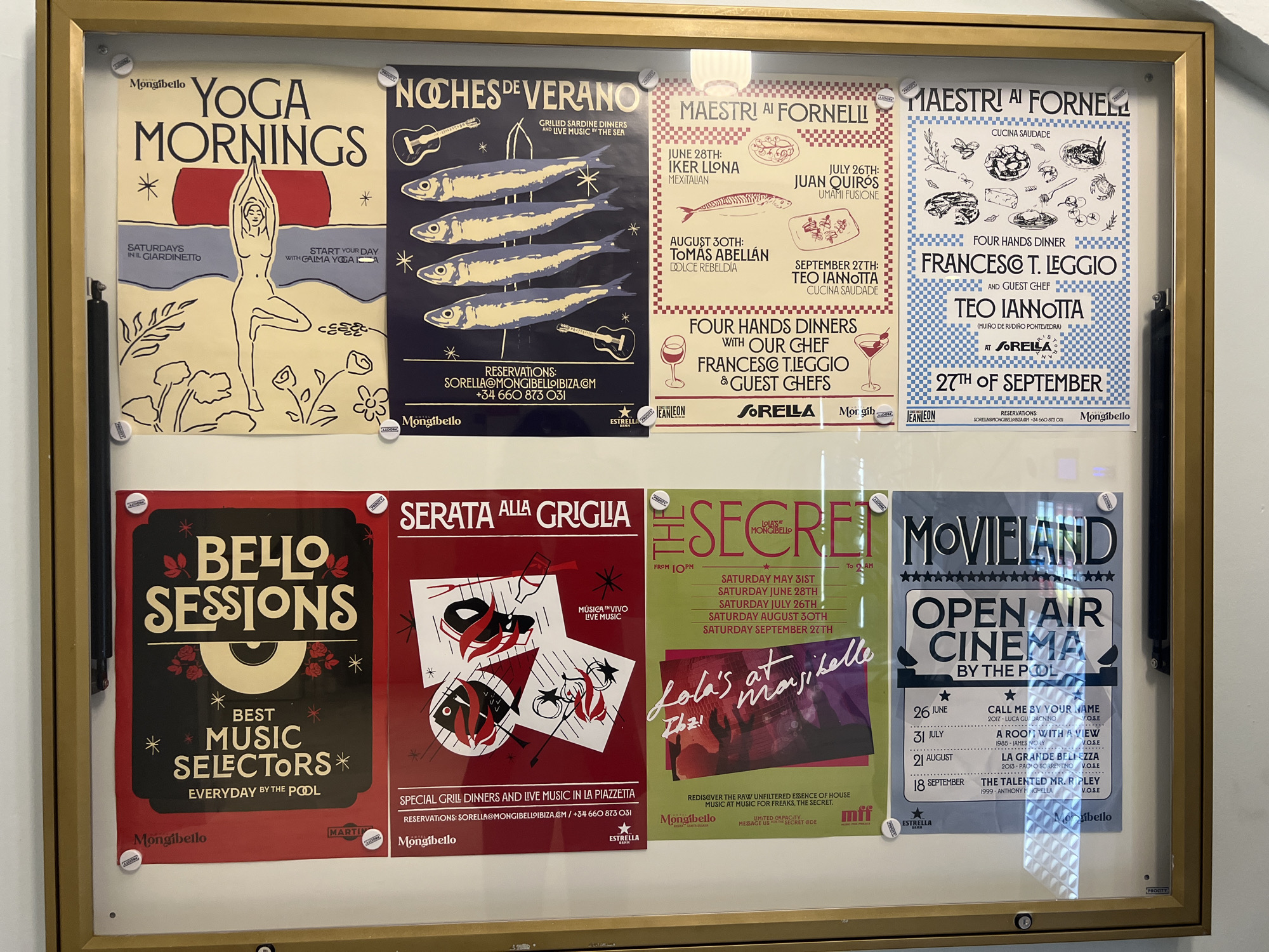 The image shows a framed display of promotional posters for events at Mongibello Ibiza, a hotel in Santa Eulària des Riu, Spain. The posters advertise yoga mornings, sardine dinners, chef collaborations ("Maestri ai Fornelli"), "Bello Sessions" music events, "Serata alla Griglia" grill dinners with live music, "The Secret" house music nights at "Lola's at Mongibello Ibiza," and an open-air cinema series ("Movieland"). Specific dates and details are visible for each event. The display is affixed to a wall with white fasteners.