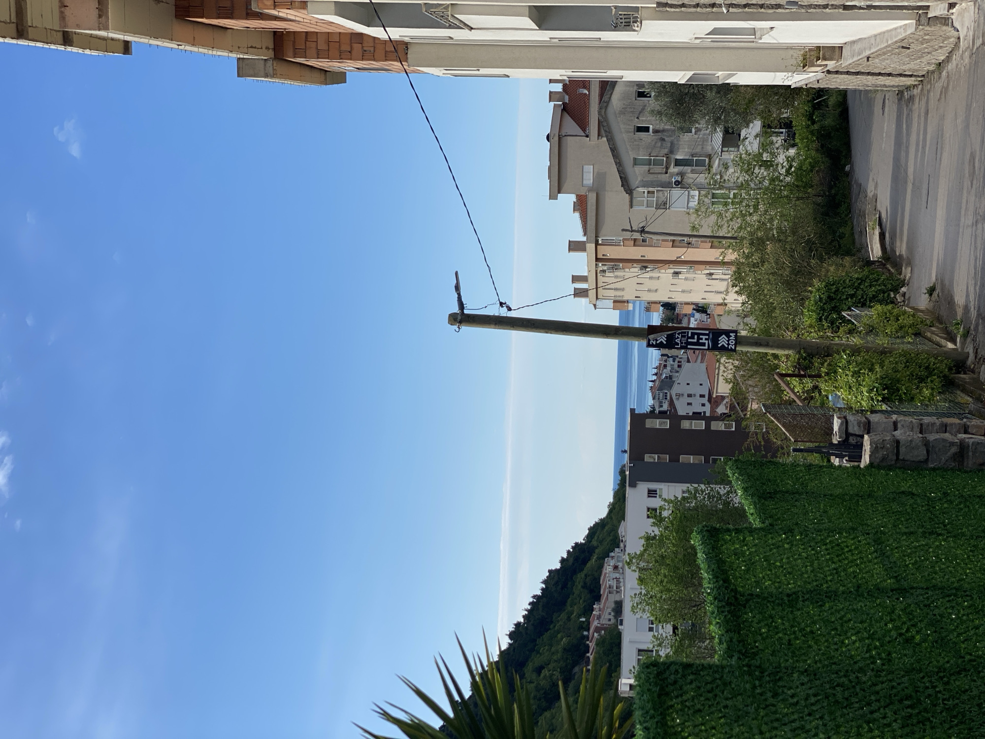 The image displays a sunny street scene in a coastal town, looking down a road towards the sea. Buildings flank both sides of the street, with a clear blue sky overhead and the sea visible in the distance.
On the left, several structures appear to be either under construction or renovation, characterized by exposed brick and concrete blocks. On the right, finished residential buildings with varying architectural styles, including white facades and darker roofs, feature multiple windows and balconies. A paved road or path winds between these buildings, descending towards the lower parts of the town.
A prominent wooden utility pole with electrical wires stands in the mid-ground, slightly to the right of the center. Attached to this pole is a black sign with white lettering. Partially legible text includes "LAZ", "N", "LH", and clearly "20M", likely indicating a distance. Further vertical text on the sign appears to be in Cyrillic script.
Beyond the immediate urban structures, a verdant hill densely covered with trees slopes down towards the horizon. The expanse of the sea, a light blue, stretches out in the background, meeting the sky. In the foreground, a vibrant green artificial hedge or fence is visible on the right, and spiky-leaved plants (possibly succulents) are in the lower left.
The scene is illuminated by bright daylight under a clear blue sky with only a few faint clouds, suggesting pleasant, sunny weather. No people or specific activities are depicted. The architectural styles, hilly landscape, and the presence of Cyrillic text are consistent with the provided location context of Budva, Montenegro.