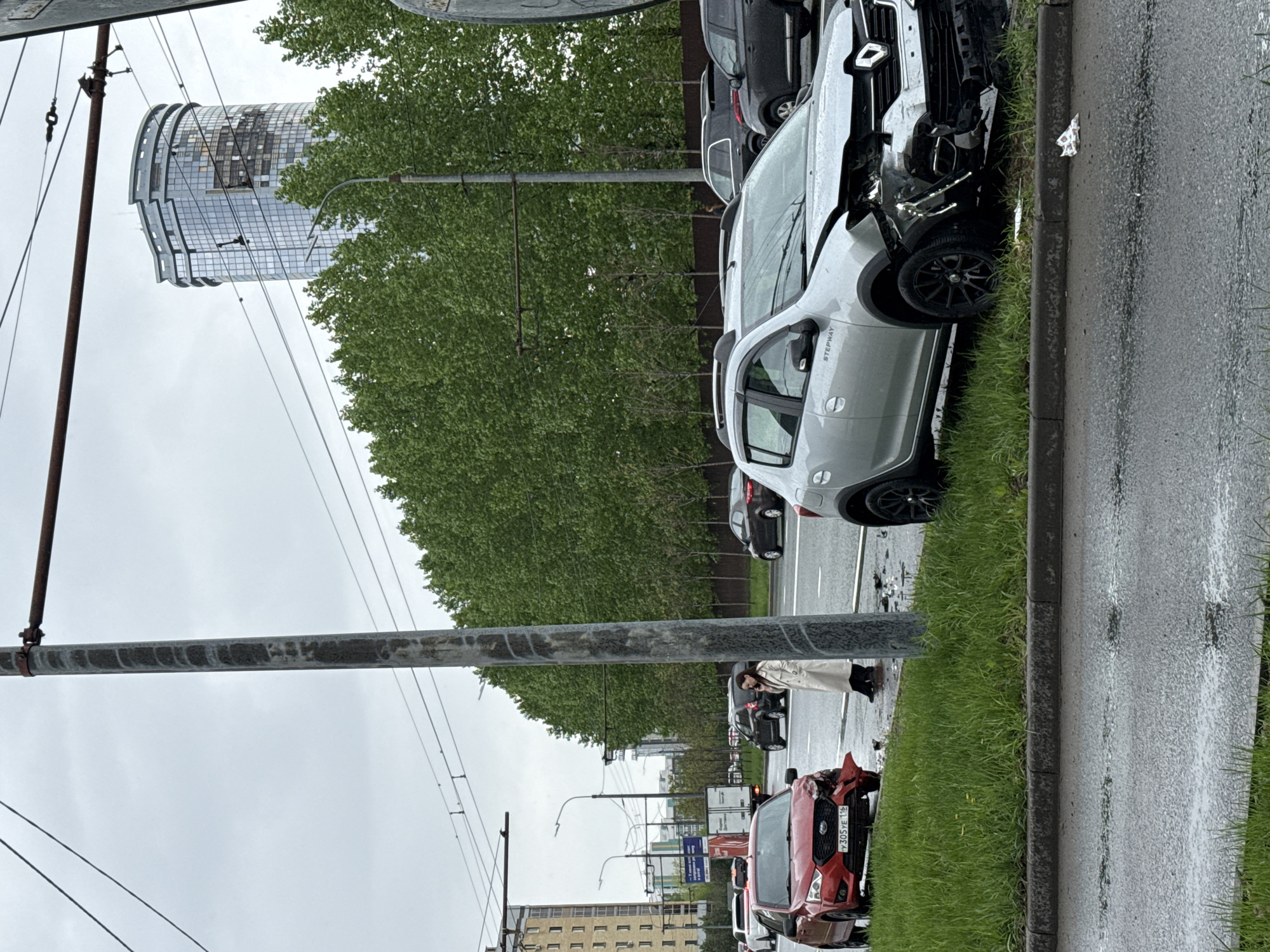 The image shows a street scene in Kazan, Russia, with two cars damaged from an accident. The first car, a silver Dacia Sandero Stepway, is partially on the curb, with the front end severely damaged. The second car, a red vehicle with the license plate "P 305 YE 116," also has visible damage. A person wearing a beige coat is standing on the sidewalk near the red car. Other vehicles are present in the background. In the distance, a distinctive cylindrical building is visible, which can be identified as a building in Kazan. The weather appears to be overcast, and the road is wet, indicating recent rain.