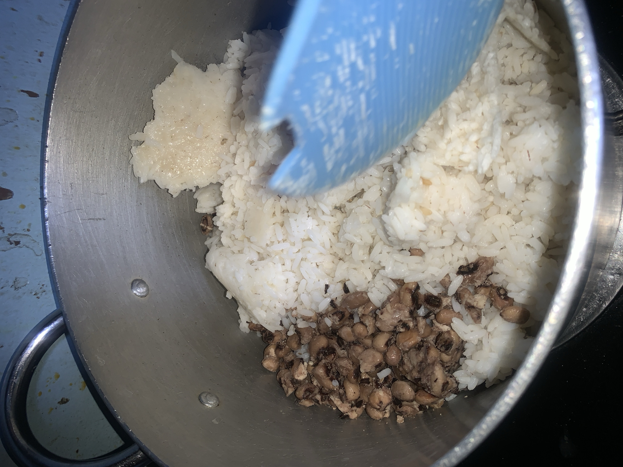 A close-up, overhead shot shows a portion of a metal pot filled with cooked food. The pot, which appears to be on a stovetop, contains a mixture of white rice and what looks like black-eyed peas with some shredded meat. The food is unevenly distributed, with a pile of rice on the right side and a mix of rice and peas on the left. A portion of a blue plastic utensil is visible on the right side of the frame, partially covering the food. The background shows the rim of the pot and a blue, speckled surface, possibly a wall or backsplash. The lighting is dim, and the image has a grainy quality. The scene appears to be indoors, in a kitchen setting.