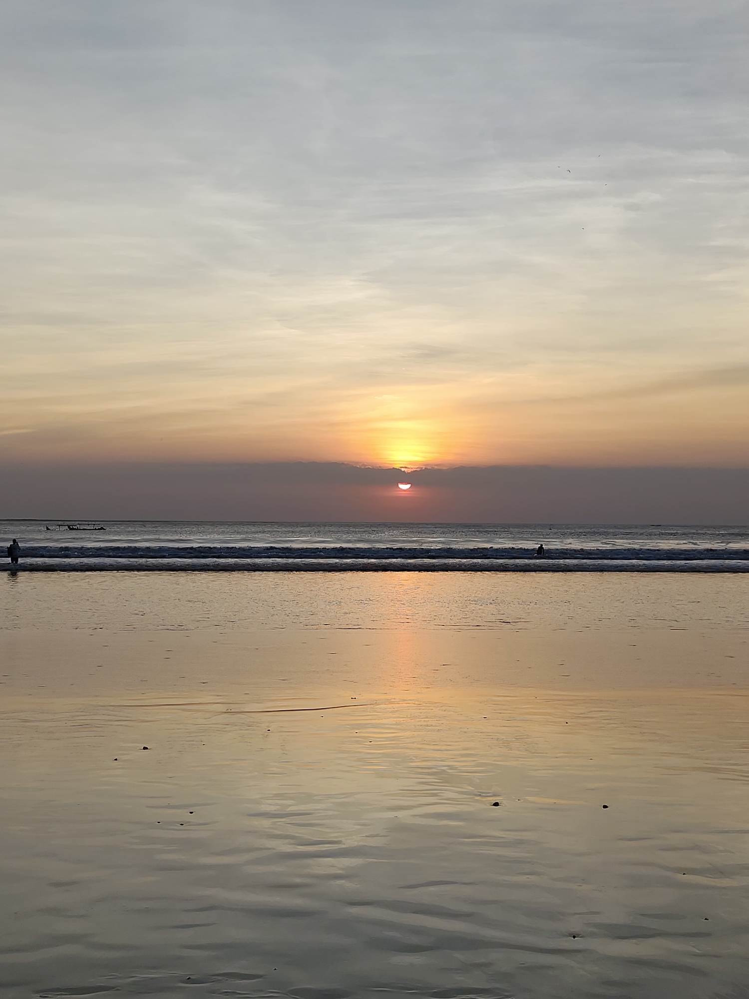 A serene beach scene unfolds at dusk, with the sun partially obscured by a thin layer of clouds near the horizon. The sky transitions from a pale gray at the top to soft hues of orange and yellow closer to the horizon, reflecting on the wet sand. Gentle waves roll towards the shore, creating a shimmering, reflective surface on the beach. A lone figure stands in the shallow water on the left side of the image, facing away from the viewer. Further out in the ocean, another solitary figure is visible, seemingly standing or sitting in the water. The overall atmosphere is one of tranquility and the quiet beauty of a sunset over the ocean.