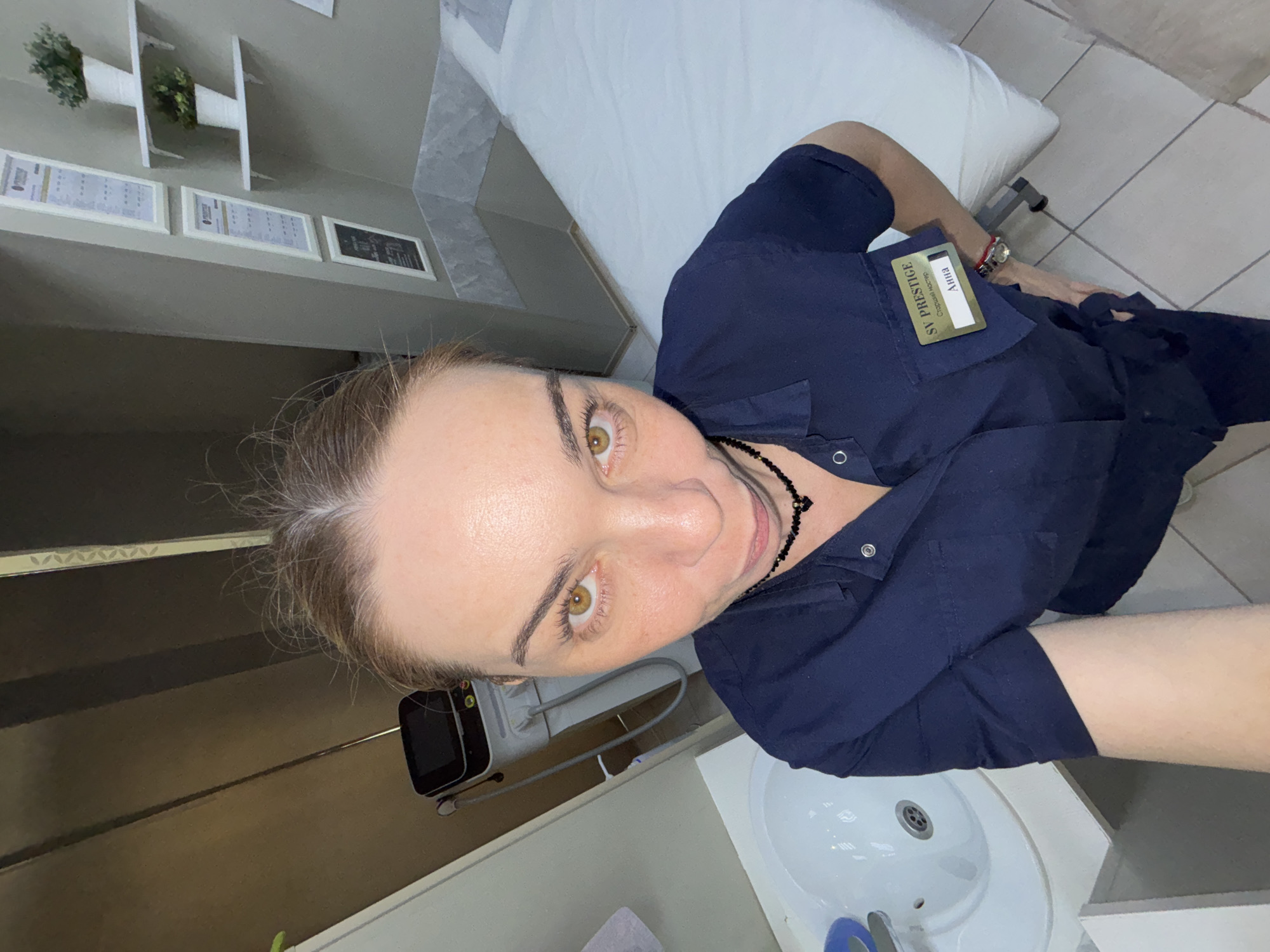 The image depicts a woman taking a selfie in what appears to be a professional setting, possibly a beauty salon, spa, or medical office. The woman has light skin, light brown hair pulled back, and striking brown eyes, looking directly at the camera with a slight smile. She is wearing a dark blue, short-sleeved uniform top with a black beaded necklace. A gold-colored name tag is pinned to her shirt, displaying "SV PRESTIGE," "Старший мастер" (Senior Master), and "АННА" (ANNA). A wristwatch with a red band is visible on her right wrist.
In the background, a white examination or spa bed with a light-colored sheet is visible to her left. To her right, a white sink and a black electronic device with a screen and cables are present. On the wall behind her, there are three framed documents or certificates, with the leftmost one showing "ПРАЙС" (PRICE), suggesting it's a service or price list. Above these documents, white shelves hold small green potted plants. The floor is covered with light-colored tiles, and the walls are light grey, with a section featuring a subtle textured pattern. The room is brightly lit, indicating an indoor environment.