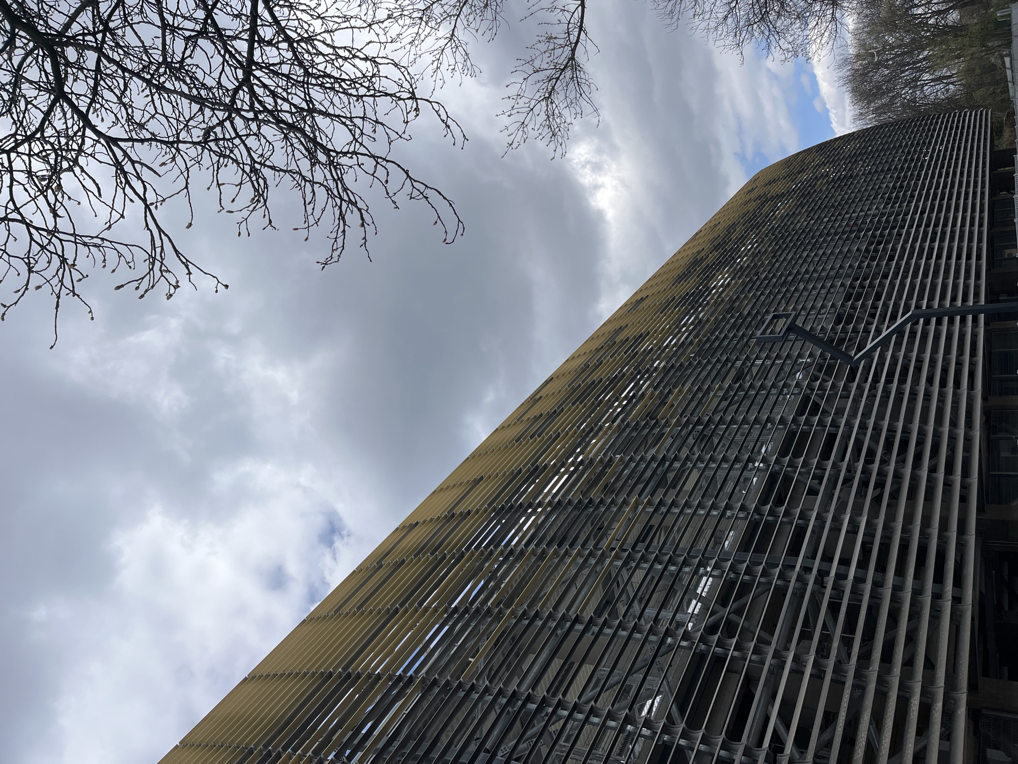 The image shows a tall structure angled against a sky filled with gray clouds. The structure is made of a lattice or grid pattern with vertical metal bars spaced closely together. The bottom left of the structure appears to have a lighter, yellowish tone compared to the darker metal on the right side. At the top left and top edge of the image, there are bare tree branches against the sky. A single, dark metal support or pole is visible on the right side of the structure. Based on its appearance, the image likely features the facade of the "St. Archangel Michael" Observation Deck in Sofia, Bulgaria.