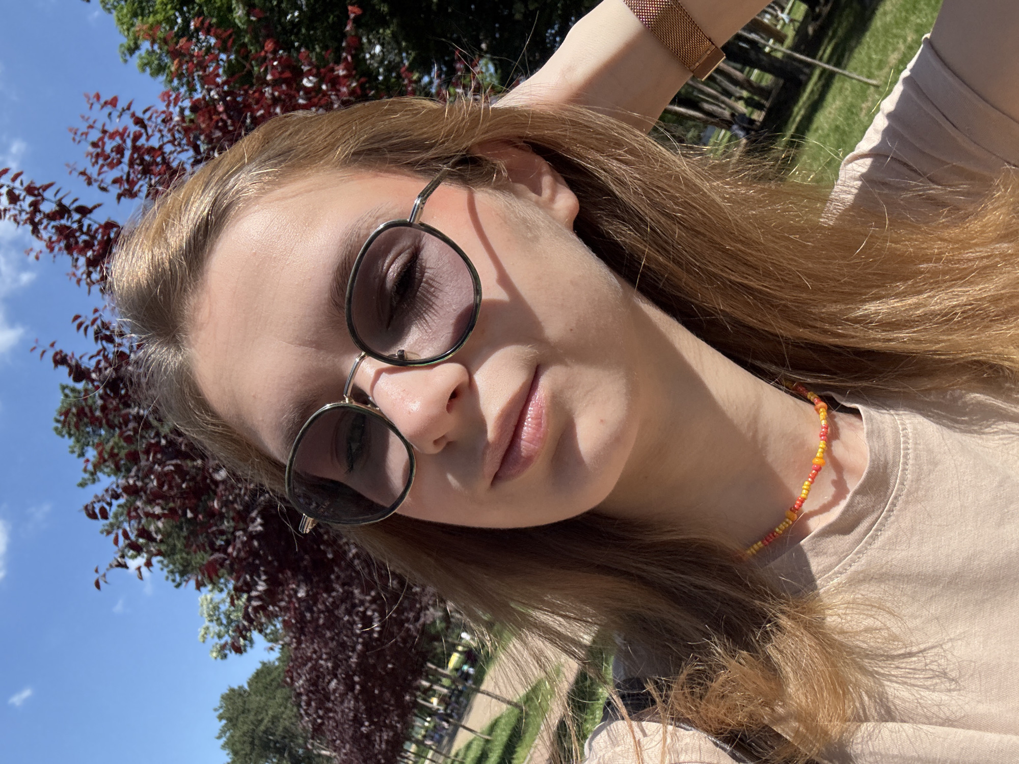 The image is a selfie of a fair-skinned person with reddish-blonde hair. They are wearing round sunglasses with a thin metal frame. The person has their hand up near their head, possibly adjusting their hair. They are wearing a beige shirt and a beaded necklace in shades of orange and yellow.
The background includes a tree with reddish leaves and a blue sky with a few white clouds. In the lower background, there are green trees and a walkway, potentially in a park or public area. Given the city context of Kragujevac, Serbia, it's plausible that this location is within one of the city's parks, such as Sumarice Memorial Park, although this cannot be confirmed with certainty.