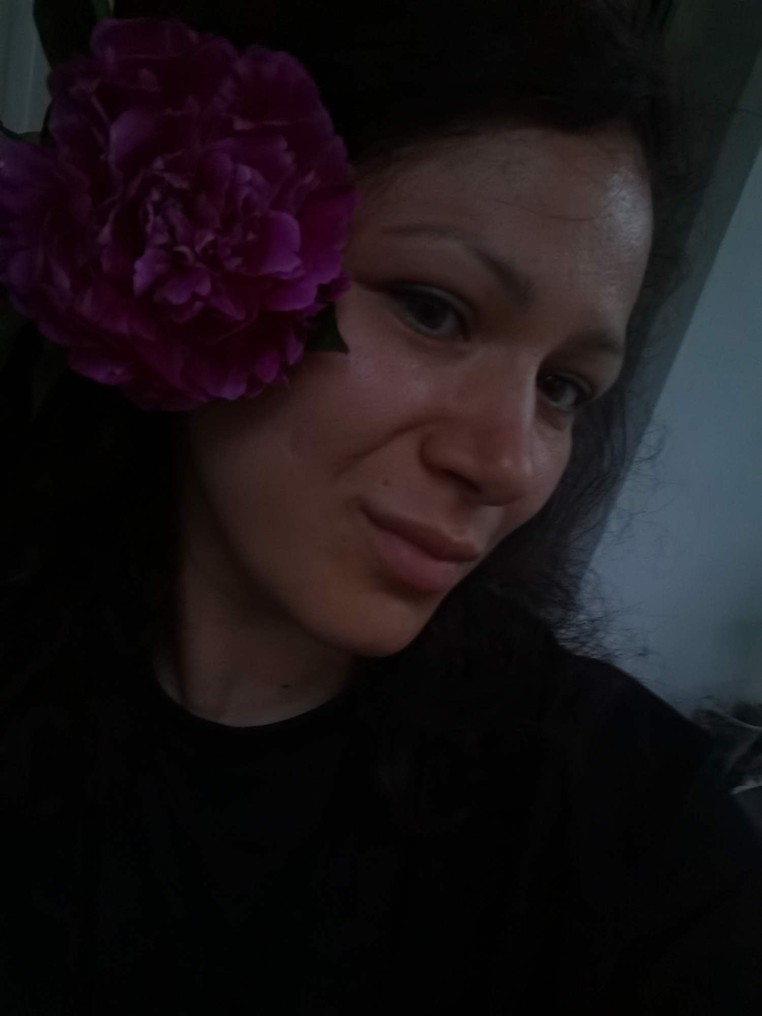 A woman with dark hair is captured in a close-up, sideways selfie. A vibrant purple flower is tucked behind her left ear, partially obscuring her face. She is wearing a black garment. The background is dark with a hint of a lighter, possibly blue or gray, wall to the right. The lighting suggests an indoor setting, possibly a home.
