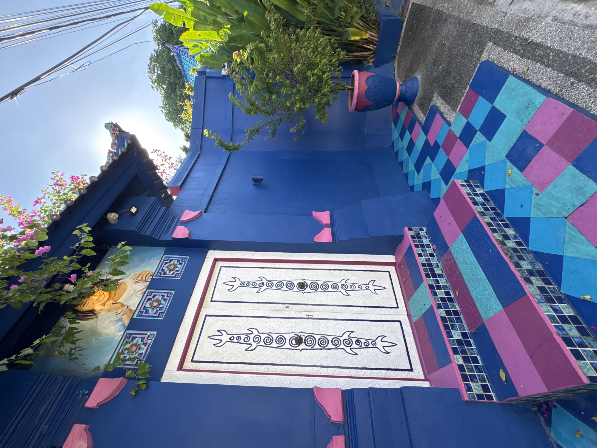The image captures an exterior scene, likely a building or a structure, in Kuta, Indonesia. The architecture displays a vivid blue color scheme complemented by pink accents. The prominent features include a set of stairs covered in a multicolored, patterned design using blues, purples, and pinks. The structure has white doors adorned with stylized dark blue designs. There is an image of a smiling girl and some decorative tiles visible on the structure. Above the structure is greenery, including a potted plant. The image is shot from a low angle, looking upwards towards the sky, and power lines are visible in the top left corner. The scene is brightly lit, suggesting a sunny day.