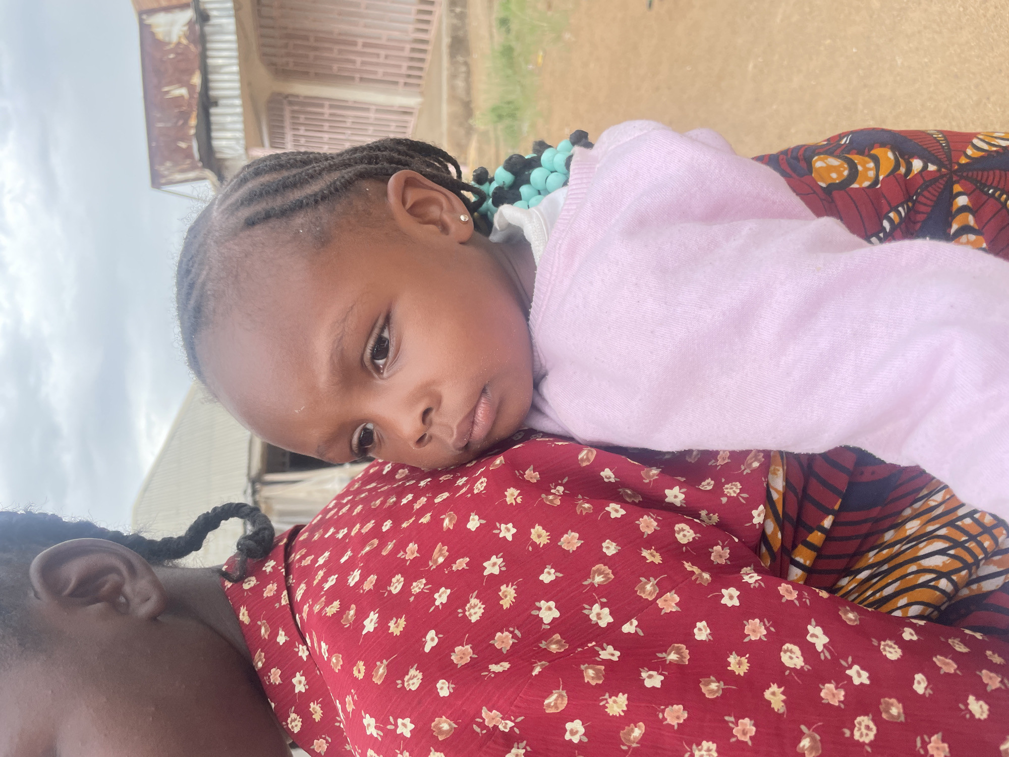 An intimate, slightly angled portrait captures a young child resting on an adult's back or shoulder in what appears to be an outdoor setting in Barakin Akawo, Nigeria. The child, likely a girl, is positioned horizontally across the adult's back, with her head and torso visible in the foreground, facing the camera. She wears a light pink long-sleeved top and has neatly braided hair. Small, sparkling stud earrings are visible in her ears. A string of light blue and black beads can be seen around her neck or shoulder, peeking out from beneath her pink top. Her expression is calm and relaxed, with a slight pout, suggesting she might be resting or feeling content.
The adult, whose ear and shoulder are visible in the lower left of the frame, is wearing a red fabric dress adorned with small, light-colored floral patterns (white, yellow, brown). There is also a glimpse of another vibrant, patterned fabric, possibly a wrapper or sling, featuring red, orange, black, and white geometric designs, visible underneath the child's body and the adult's red dress. The adult also has braided hair.
The background shows an overcast sky, suggesting a cloudy daytime. Hints of rustic structures or buildings, including corrugated metal and what looks like brick or concrete walls, are visible, typical of a residential or rural area. There is no visible text in the image.