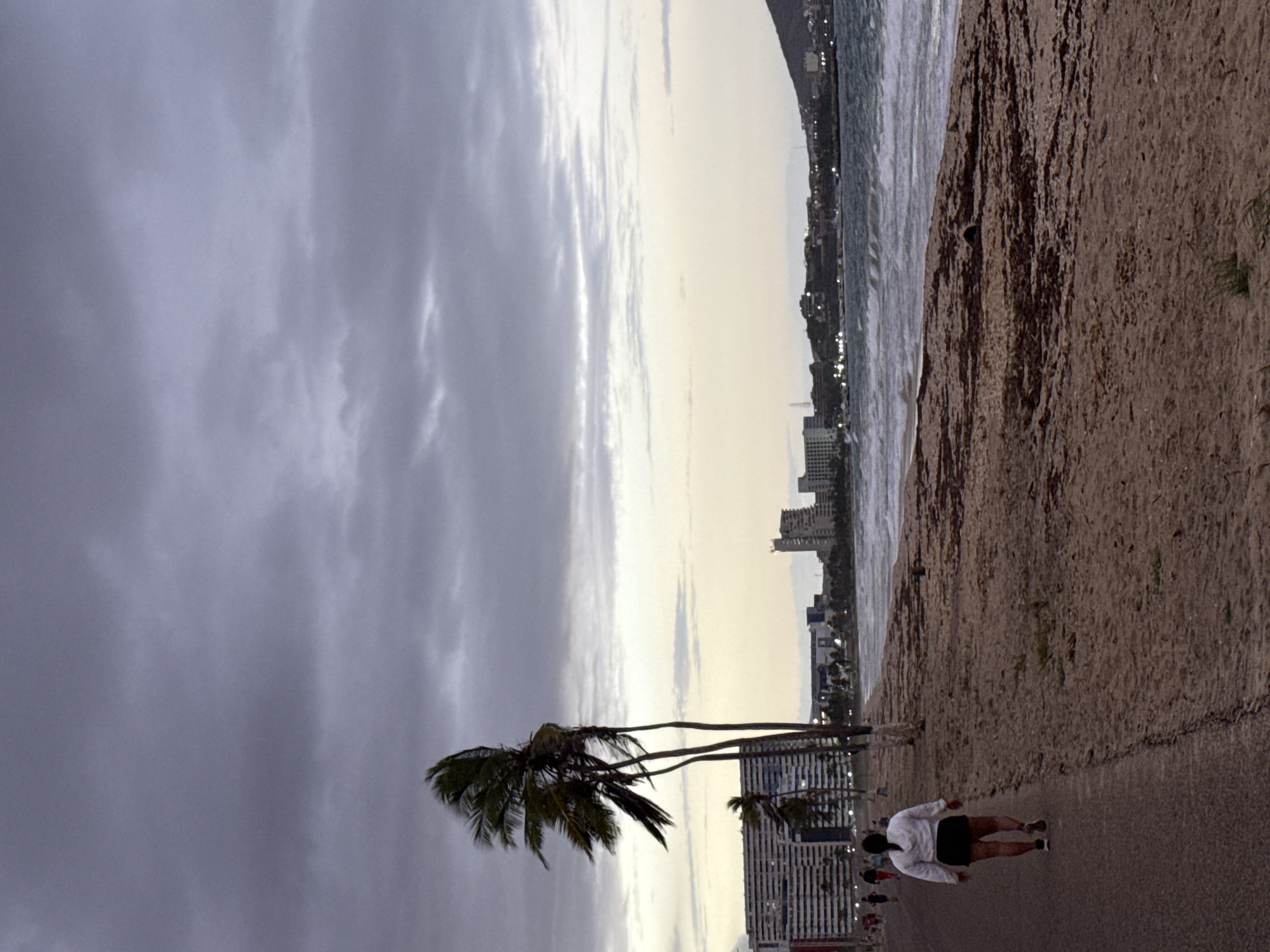 The image depicts a coastal scene in Porlamar, Venezuela, during either dusk or dawn. The view is oriented along a promenade running parallel to the ocean.
In the foreground, a paved pathway occupies the left side of the frame, where a person is walking away from the viewer. This individual is wearing a white long-sleeved top, dark shorts, and has their hair pulled back. To their right, a sandy beach transitions into the ocean, with gentle waves visible near the shore. The sand appears textured and slightly rocky in areas.
Further into the mid-ground, two tall palm trees with green fronds stand out against the sky, adding a tropical touch to the landscape. Beyond the beach and ocean, a city skyline stretches across the horizon, with numerous buildings and faint, twinkling lights suggesting the early evening or pre-dawn hours. The buildings appear to be part of a coastal urban development.
The sky above is heavily overcast, filled with various shades of gray clouds, creating a subdued atmosphere. Towards the horizon, the clouds lighten, revealing a strip of brighter sky that indicates the time of day. No discernible text is visible in the image. The overall impression is one of peaceful coastal activity, possibly a walk or jog along the waterfront.