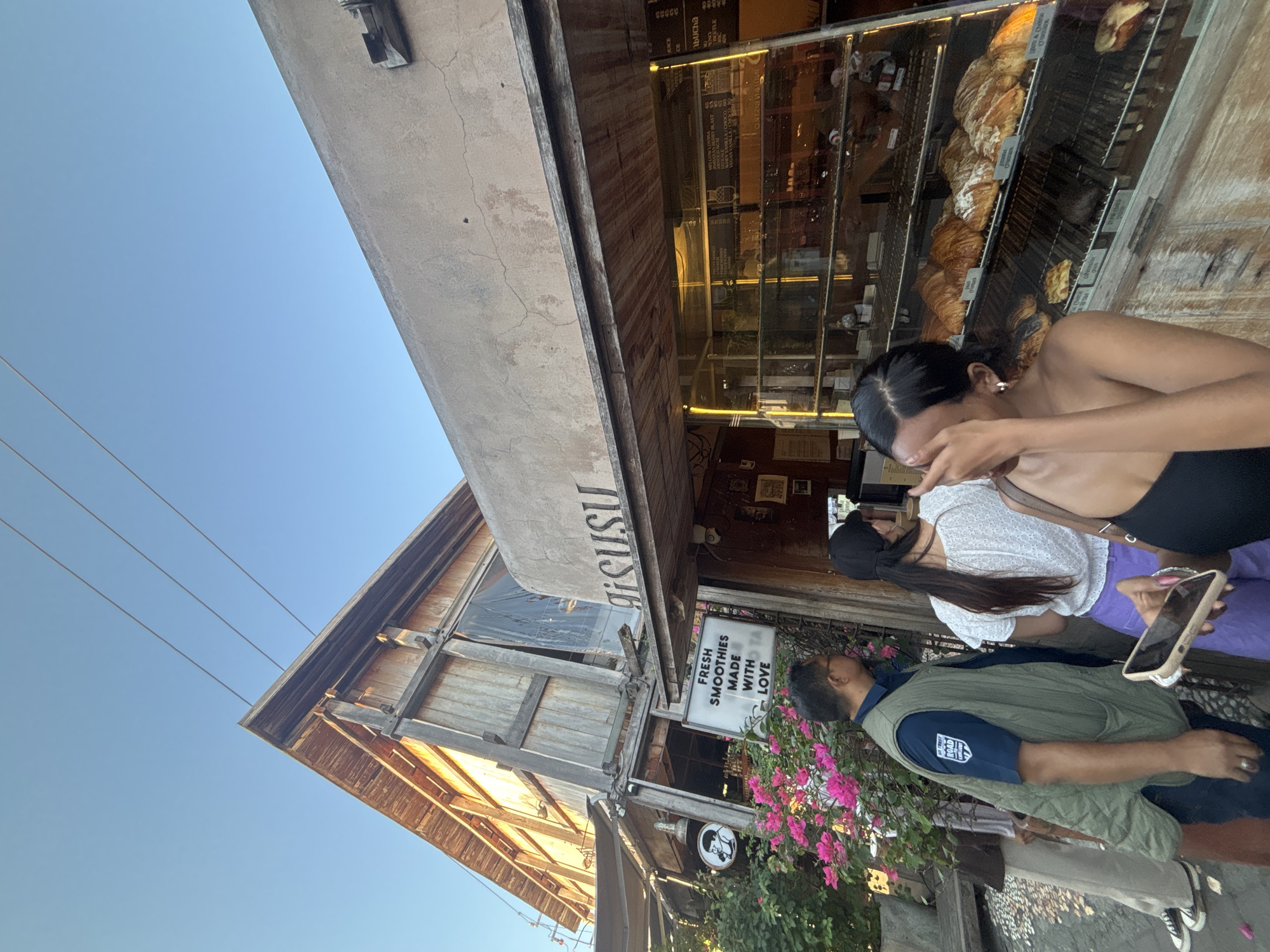 The image depicts the exterior of a cafe in North Kuta, Indonesia. The cafe has a rustic, unfinished wooden structure with a concrete facade. A sign above a display window reads "gisusu". Inside the window, shelves are filled with croissants and other baked goods. A menu board is visible behind the display. In the foreground, several people are standing. To the left, a man in a green vest and a blue shirt with a patch on the sleeve stands near a sign that reads "FRESH SMOOTHIES MADE WITH LOVE" and a bush with pink flowers. To the right, two women are partially visible. One woman wears a white top and purple pants, and the other, in the immediate foreground, is wearing a black top and covers her face with her hand. The sky is clear and blue, and power lines run across the upper portion of the image.