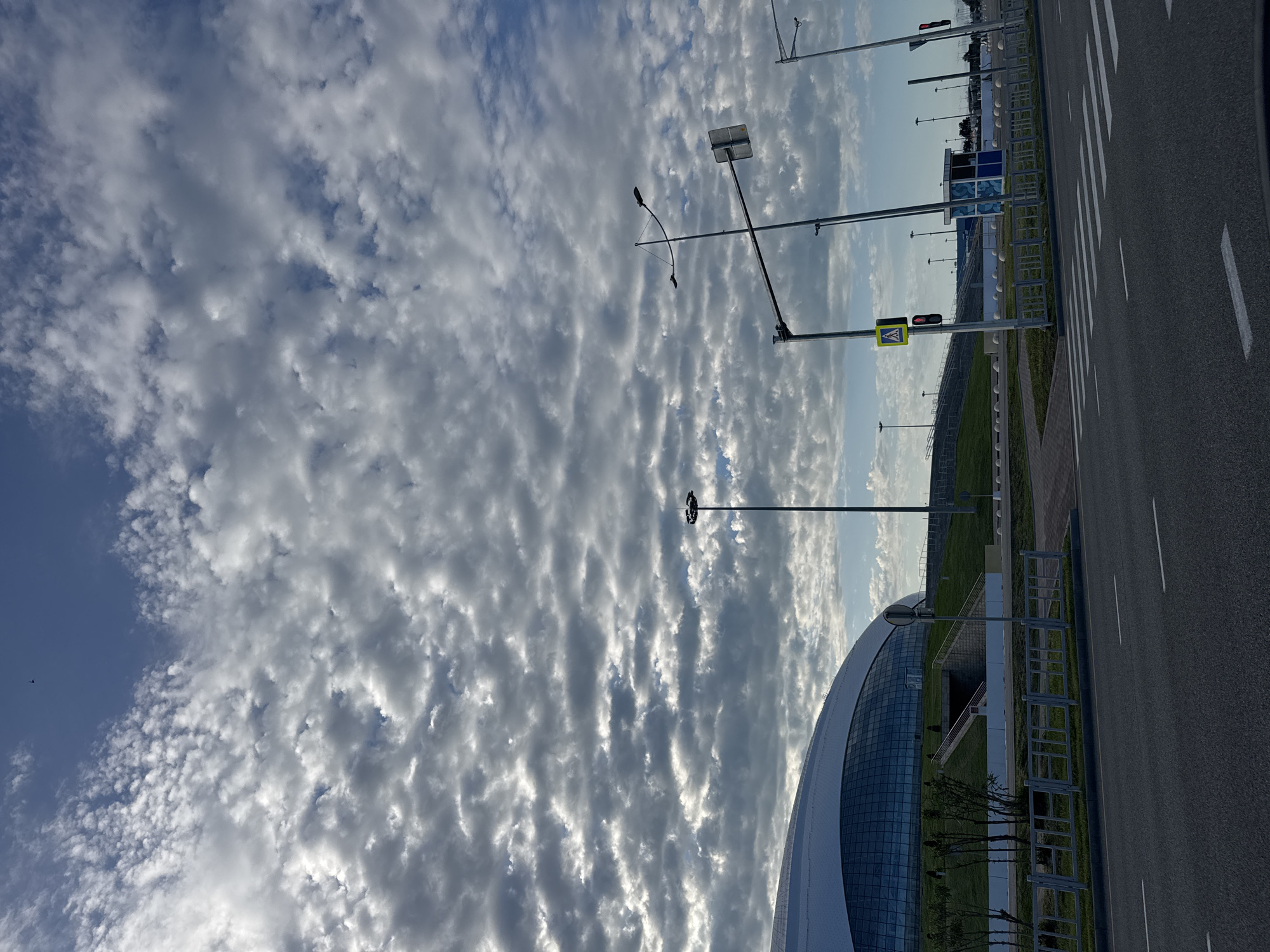 The image displays an expansive outdoor scene, dominated by a dynamic sky and modern infrastructure. The upper left and central portions of the frame are filled with a striking pattern of altocumulus clouds, presenting a rippled or patchy appearance against a backdrop of blue sky. Small glimpses of direct sunlight illuminate the edges of some clouds. A tiny dark speck, possibly a bird or distant aircraft, is visible against a clear patch of blue sky in the upper left.
The lower and right sections of the image feature a paved road with distinct white markings, including solid lines and the broad stripes of a pedestrian crossing. Several tall, dark lamp posts or utility poles are scattered across the scene. One prominent pole on the right side of the road features a yellow diamond-shaped sign with a black pictogram of a person walking, indicating a pedestrian crossing. Below this, a traffic light is visible, showing a red signal.
To the left of the road, a strip of neatly maintained green grass separates the thoroughfare from a large, modern building. This building exhibits a curved architectural design with a reflective glass facade, suggesting a contemporary public or commercial structure. Fences and barriers are present along the roadside and near the building, along with some small trees or bushes. The scene is devoid of people or moving vehicles, creating a quiet and somewhat serene atmosphere. The overall impression is that of a well-maintained, potentially newly developed area, under bright daylight conditions.