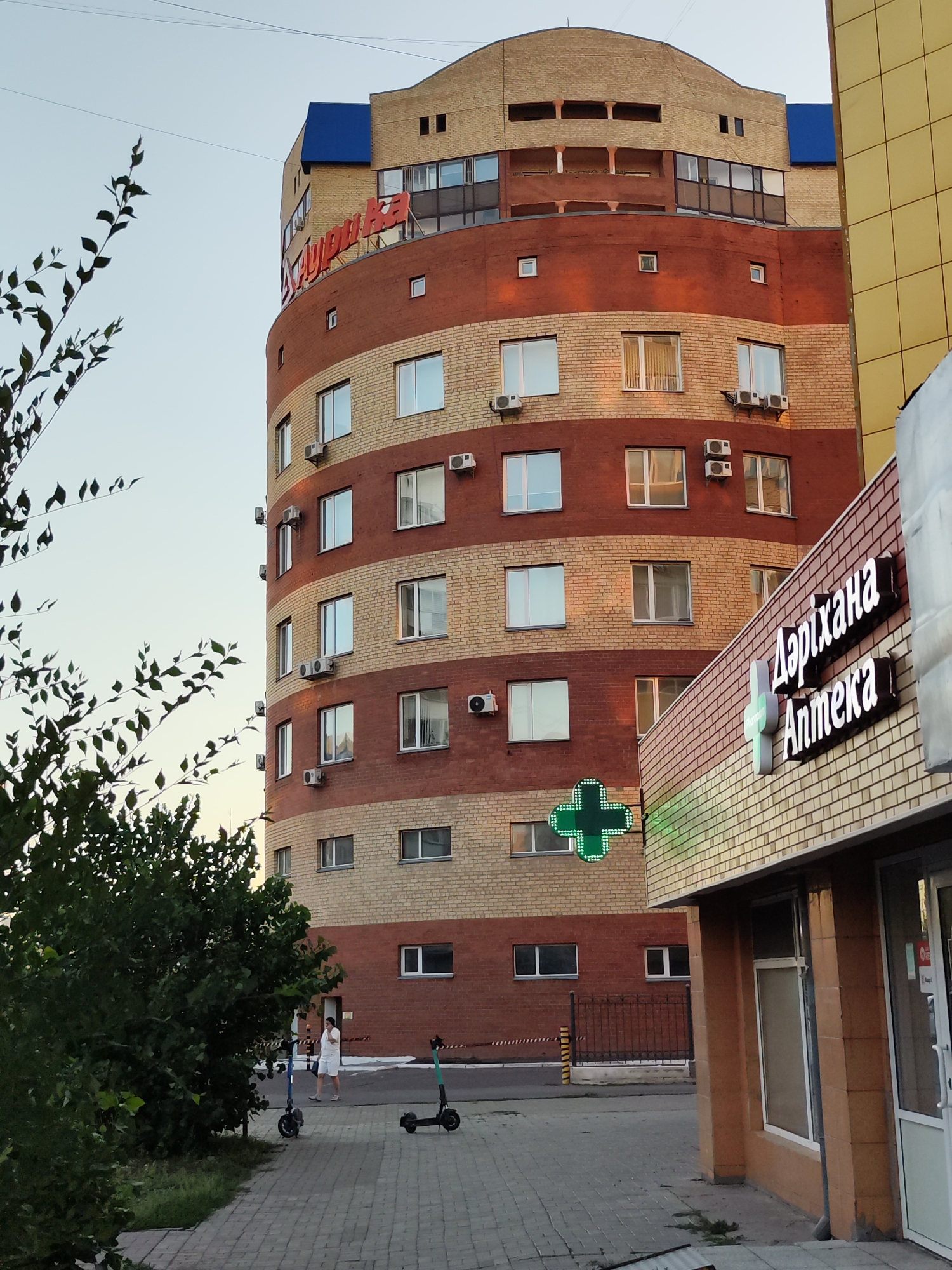 The image shows a curved, multi-story building with alternating bands of red and beige bricks, located in Astana, Kazakhstan. A prominent red sign with Cyrillic lettering is visible at the top of the building. To the right, another building with a yellow tiled facade is partially visible, and a pharmacy with a neon green cross and signage is situated at street level. In the foreground, a sidewalk with cobblestone paving is present, with a person walking and two electric scooters parked nearby. Green foliage from trees borders the left side of the image. The sky is clear with a pale blue hue.