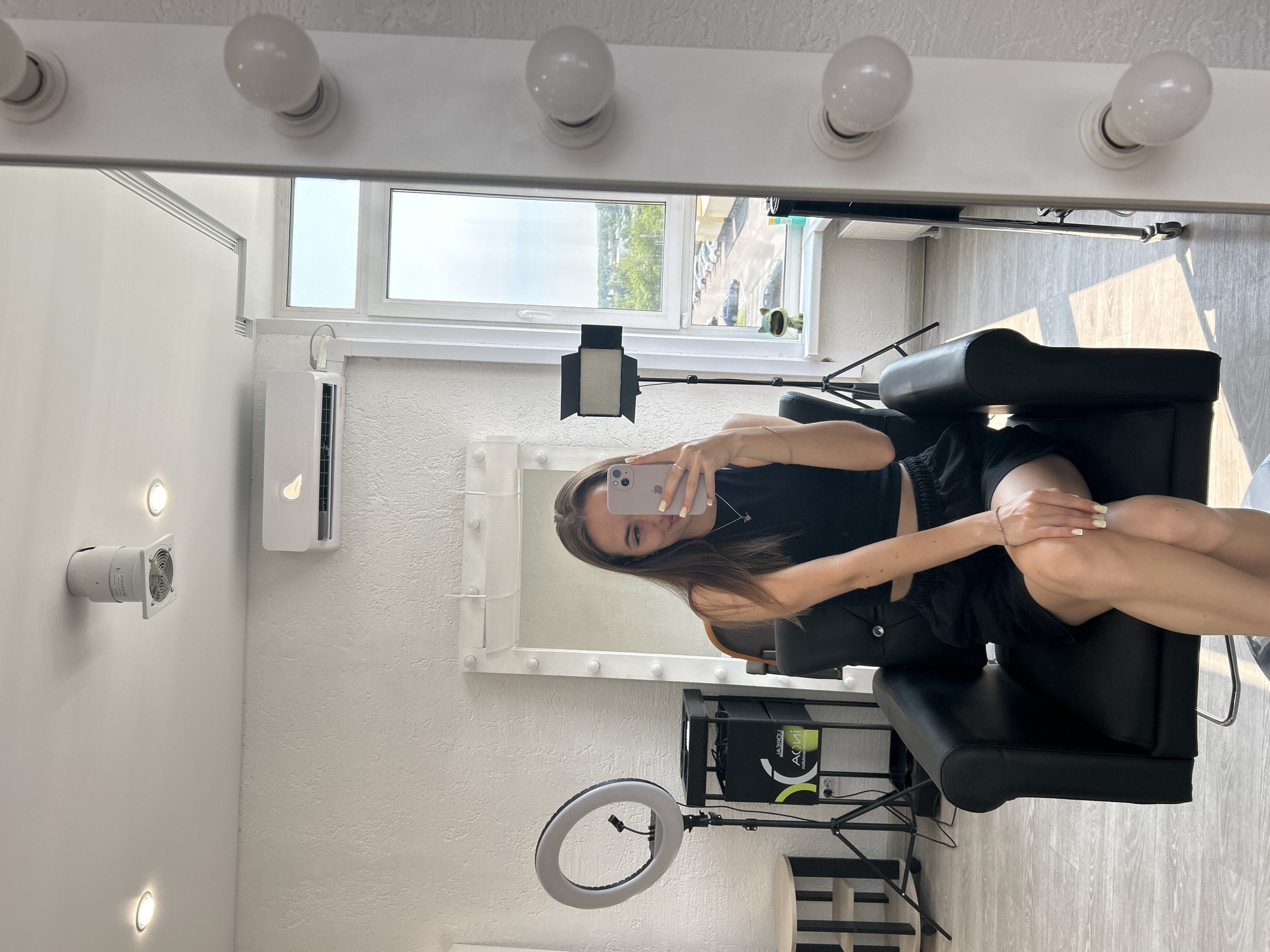 A young woman with long brown hair is taking a selfie in a brightly lit room, likely a hair salon or dressing room. She is wearing a black crop top and black shorts. She is sitting in a black leather chair and holding a smartphone in her right hand. The room has a large vanity mirror with light bulbs around the edge, a ring light on a stand, and another professional light on a tripod. A window offers a view of a street with parked cars and some greenery. The walls are white and textured, and the floor is light-colored wood. An air conditioning unit is visible on the wall.