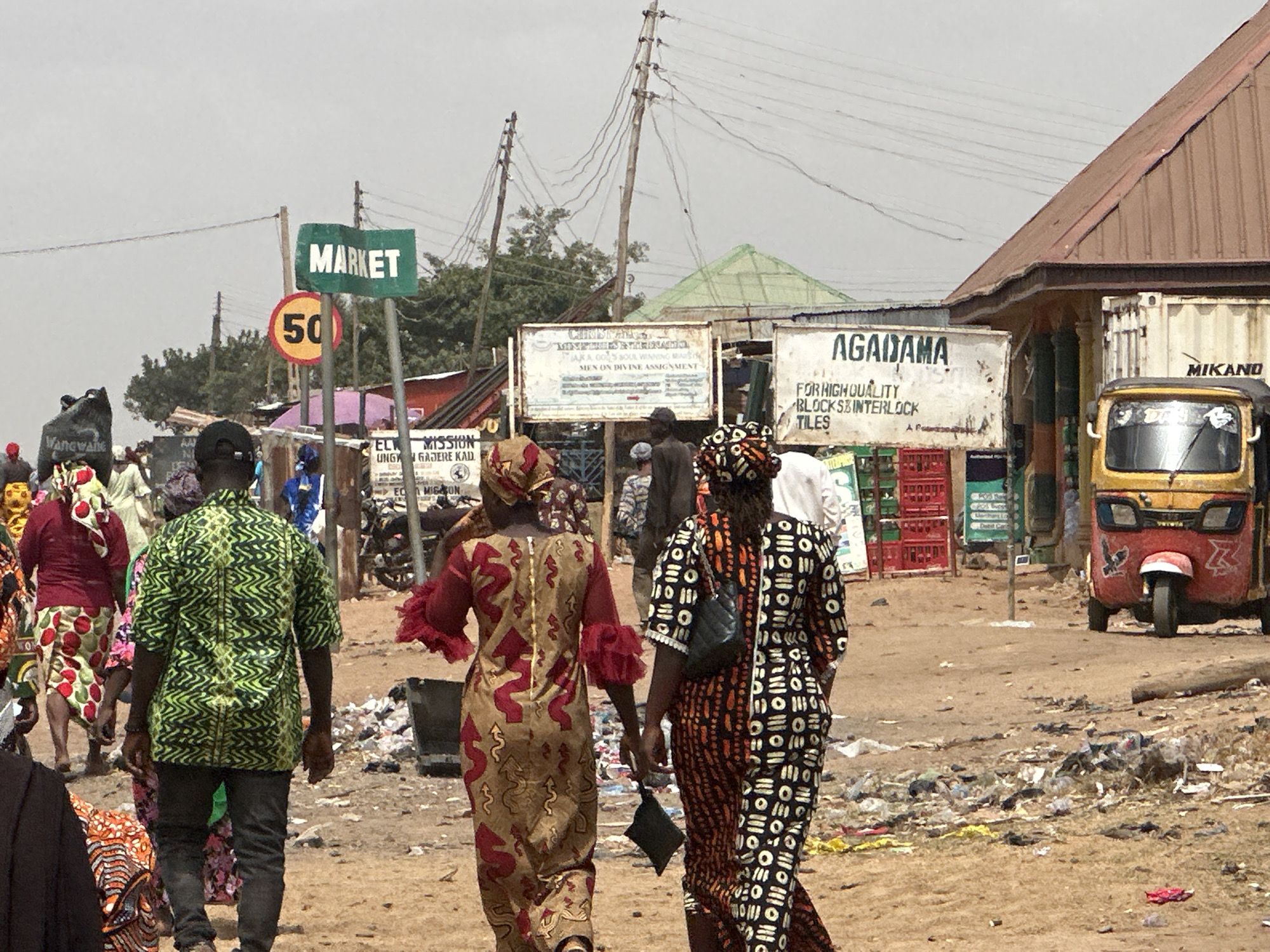 The image captures a street scene in what appears to be a developing area. The scene is populated by people wearing traditional African garments, walking on a dirt road scattered with trash. In the background, there are various signs, including one pointing towards "MARKET" and a "50" speed limit sign. Additionally, there's a sign advertising "AGADAMA FOR HIGH QUALITY BLOCKS & INTERLOCK TILES." An auto rickshaw (tuk-tuk) is parked on the side. Power lines crisscross overhead. The sky is overcast, and the general environment suggests a bustling market area in a city.