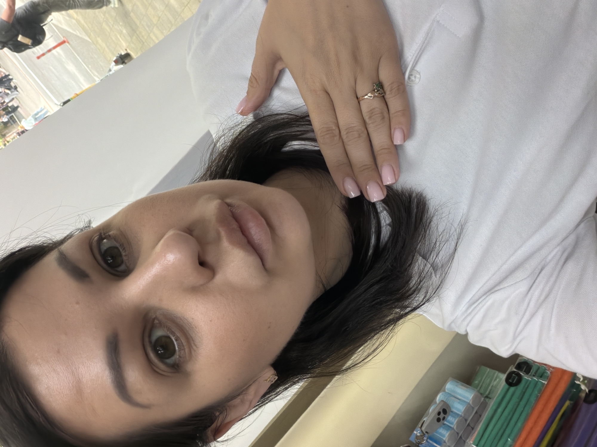 This is a medium shot, taken from a slightly high angle, of a woman posing for a selfie. The woman has dark brown hair and light brown eyes. She is wearing a white polo shirt and a small gold earring in her left ear. Her right hand is resting on her chest, and she is wearing a gold ring with a green stone on her right ring finger. Her fingernails are painted a light pink color. The background is out of focus but appears to be an indoor setting with shelves of colorful items to her left, and a blurry outdoor scene with people and a street to her right. The woman's facial expression is neutral. There is no specific indication of the location being Sochi, Russia.
