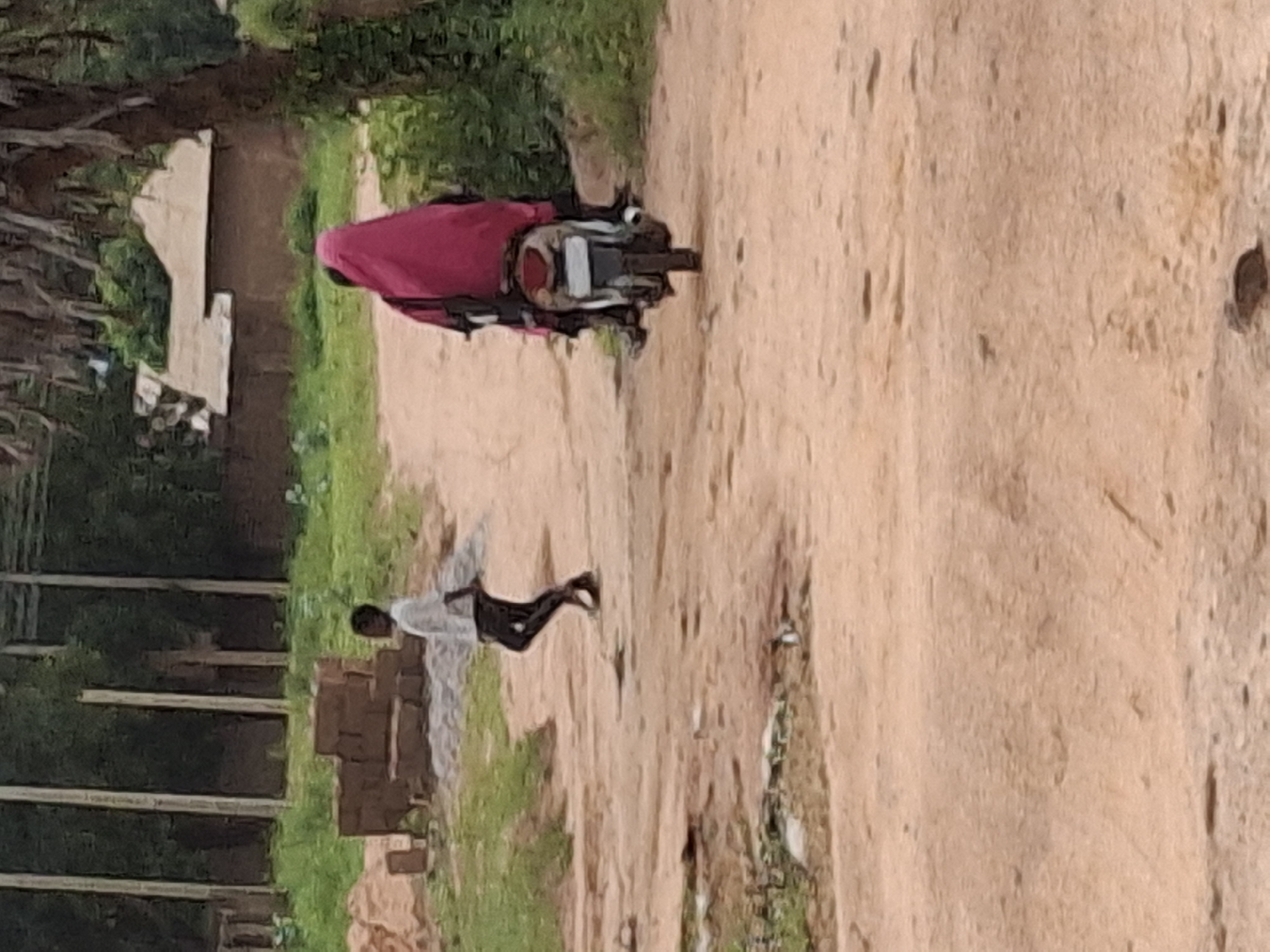 In Tempure, Nigeria, a dirt road is visible with scattered debris. To the left, a pile of bricks is stacked. In the middle ground, a young boy in a white shirt and dark pants walks on the road, facing away from the camera. Further back on the right side of the road, a person wearing a bright pink headscarf is seated on a motorcycle, also facing away from the camera. The background features trees and a building with a thatched roof.