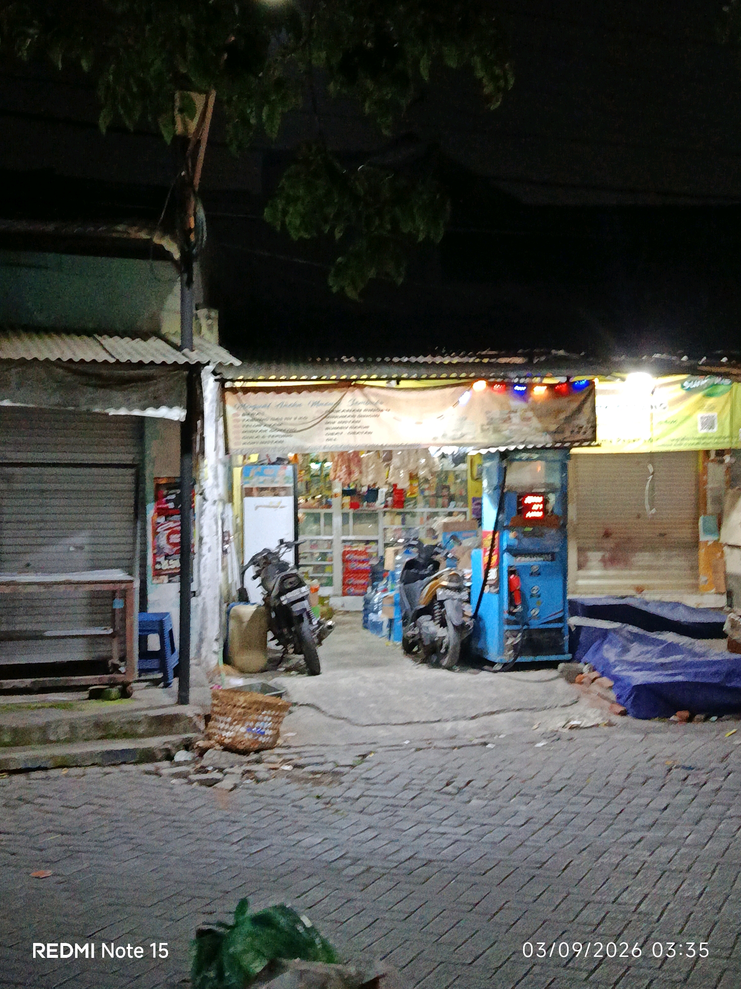 The image depicts a street scene at night in Keboansikep, Indonesia, featuring a small convenience store or warung with a blue gas pump. The store is illuminated by overhead lights, some of which are colored red and blue. Shelves stocked with various goods are visible inside. In front of the store, two motorcycles are parked, one black and white, and another partially obscured. A woven basket sits on the cobblestone ground near the motorcycles. To the left, a metal-framed shelf unit is placed in front of a closed roll-up door. A blue plastic stool is positioned next to the shelf unit. The pavement is made of interlocking bricks, and overhead foliage partially obscures the dark night sky. The timestamp "03/09/2026 03:35" is visible in the lower right corner of the image, along with the text "REDMI Note 15" in the lower left.