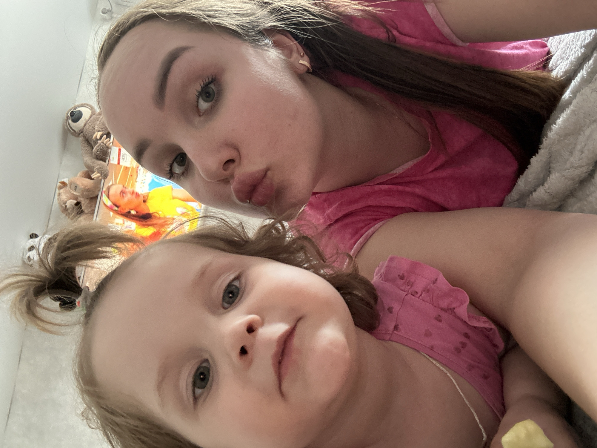 Two people, likely a mother and daughter, are posing for a selfie. Both are wearing pink shirts. The woman has long brown hair and is making a kissing face. The child has light brown hair styled in a ponytail. A teddy bear and a magazine are visible in the background. The location and city are unknown.