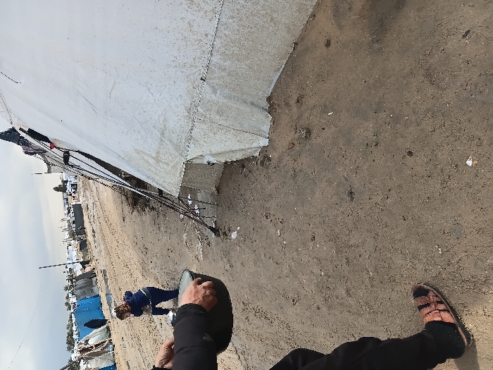 A low-angle shot shows the exterior of a large white tent structure. Ropes and stakes anchor the tent to the sandy ground. In the distance, blue tents and buildings are visible under a cloudy sky. The viewer's perspective includes a person's legs in black pants, sandals on one foot, and a hand holding a dark object, possibly a helmet. A woman in a blue outfit is partially visible near the distant tents.