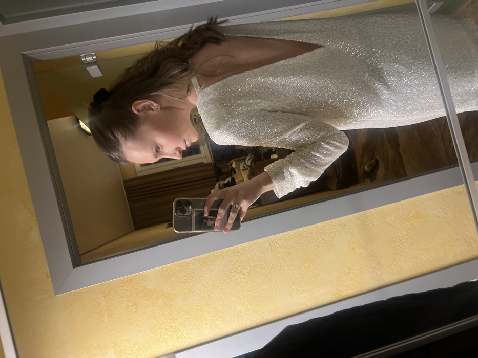 A mirror selfie captures a woman in a shimmering white dress with a low back and long sleeves. She holds a phone with a clear case, partially obscuring her face. The reflection shows a hotel room setting with dark wood tones and tan walls. The mirror has a gray frame, and the wall behind it is painted a textured yellow. A dark garment is visible at the bottom of the frame.