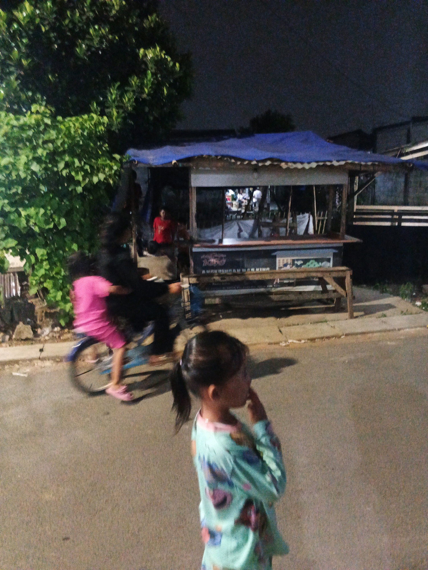 In Kadu, Indonesia, at night, a young girl in a blue and green long-sleeved shirt stands in the foreground, facing right with her hand near her mouth. Behind her, two people are on a bicycle. One person, wearing a pink shirt and shorts, is on the seat, and another person in black is behind them, pedaling. The bicycle is in motion, indicated by the blur of its wheels. In the background, a food stall with a blue tarp roof is illuminated. Several people are visible inside and around the stall, some appearing to be customers or vendors. Trees and some buildings are also present in the background, with the sky dark overhead. The scene is captured on a paved road with some debris on the side.